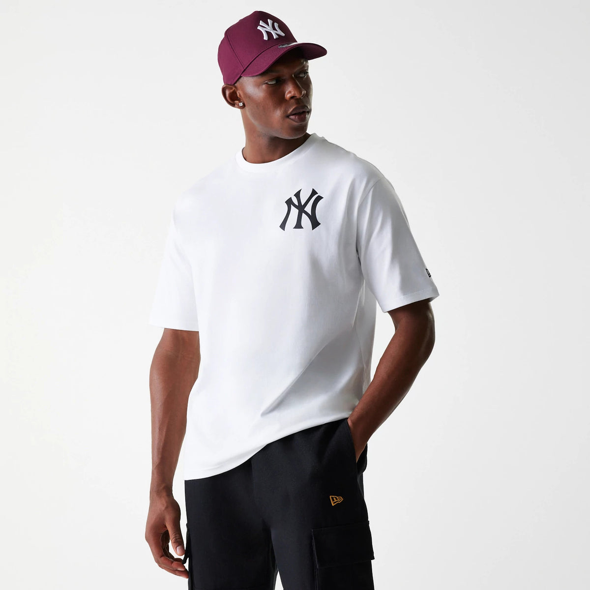 New Era New York Yankees MLB Baseball Graphic White Oversized T-Shirt, on-model view showing a white shirt with a black New York Yankees logo on the left chest, worn by a man in black shorts with a maroon cap featuring a raised logo.