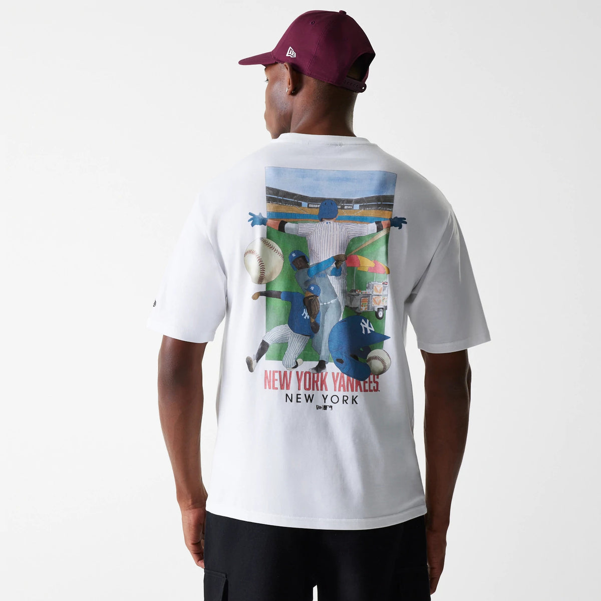 New Era New York Yankees MLB Baseball Graphic White Oversized T-Shirt, rear view showcasing an illustrated baseball scene with players and balls, below text reading 'New York Yankees New York', on-model with a burgundy cap.