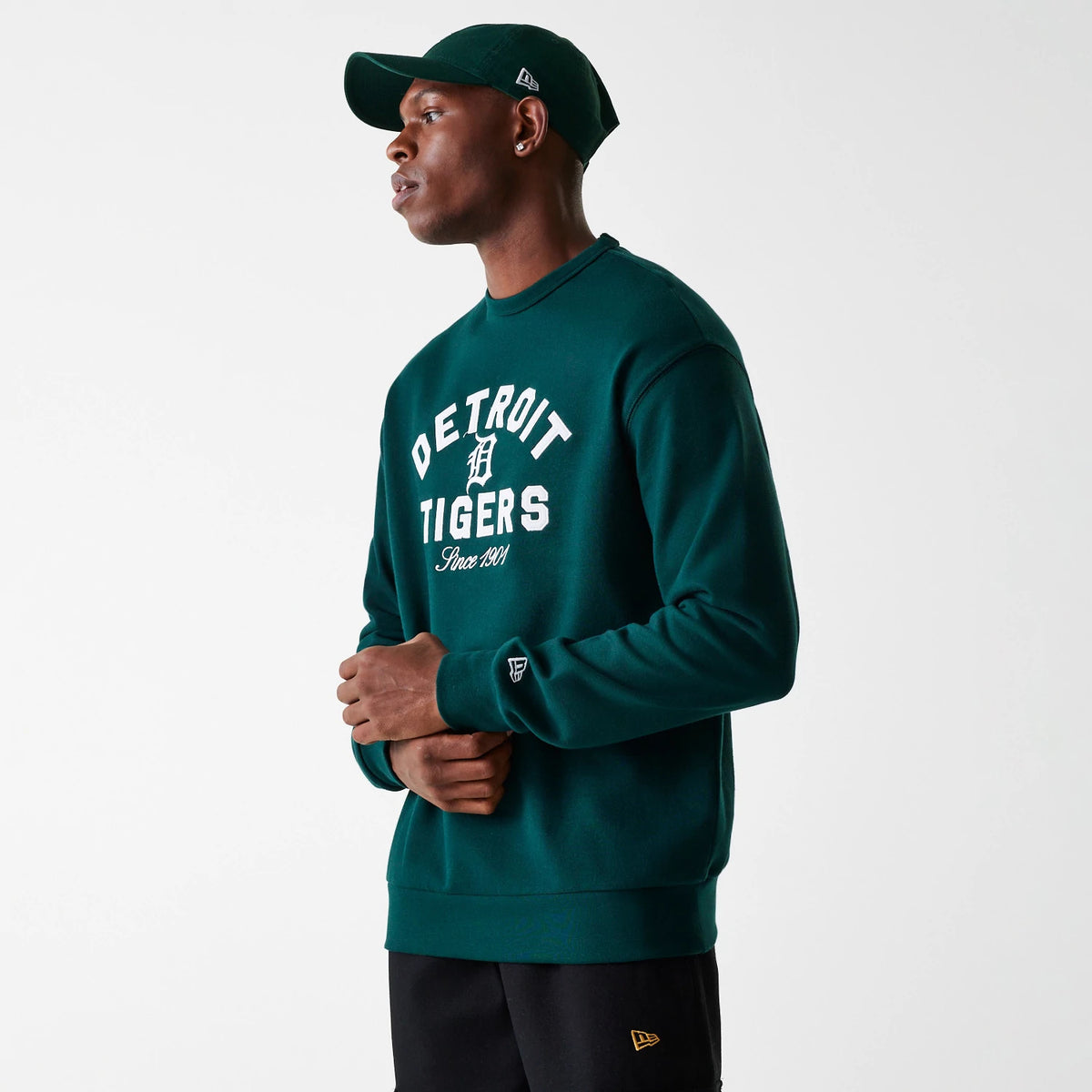 New Era Detroit Tigers MLB Heritage Dark Green Oversized Crew Neck Sweater, on a model wearing a matching cap, showing large white embroidered Detroit Tigers lettering across the chest and a New Era flag logo on the left sleeve cuff.