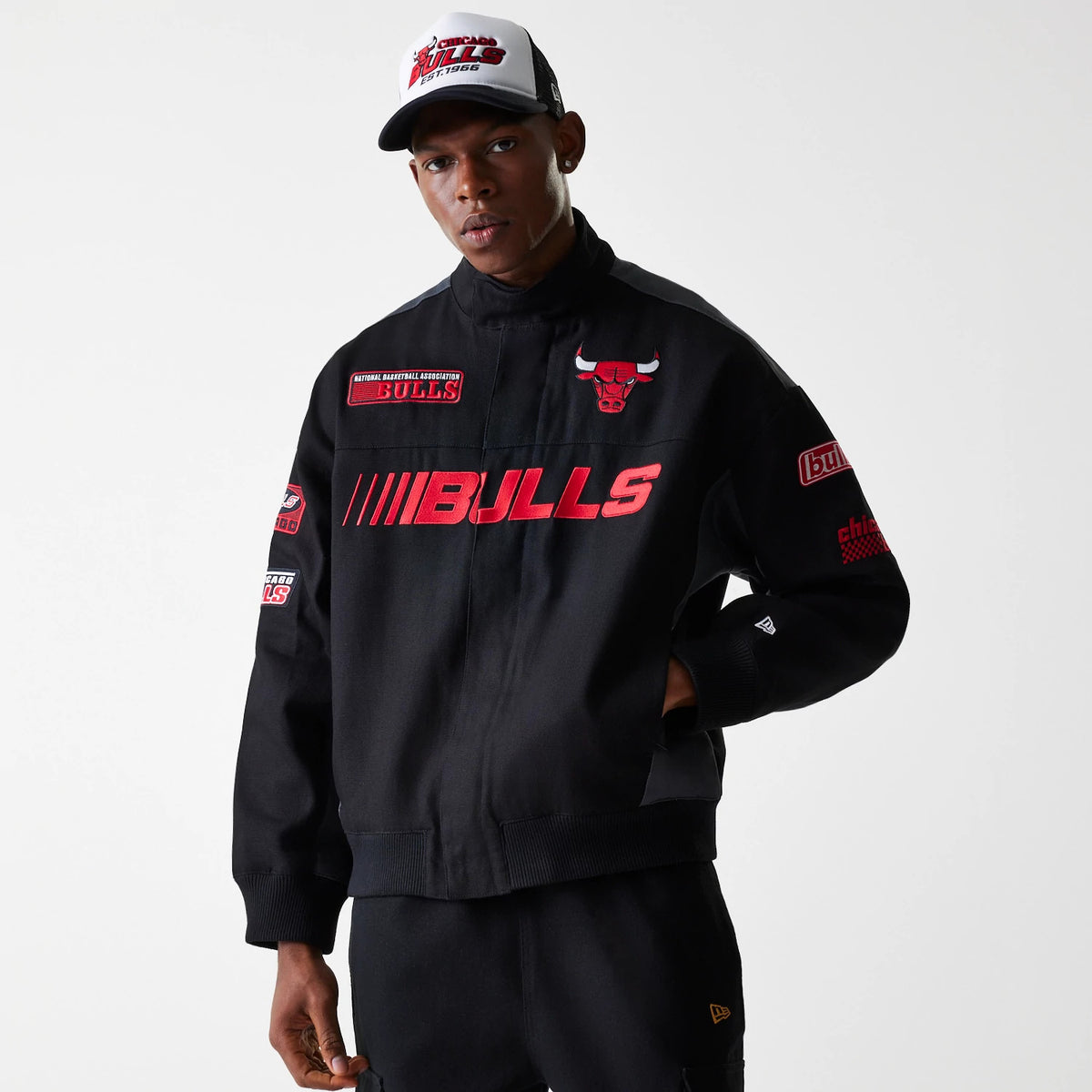 New Era Chicago Bulls NBA Black Motorsports Jacket, front view showing the black jacket with red BULLS lettering across the chest, a red and white embroidered bull logo on the left chest, and various patches on the sleeves.
