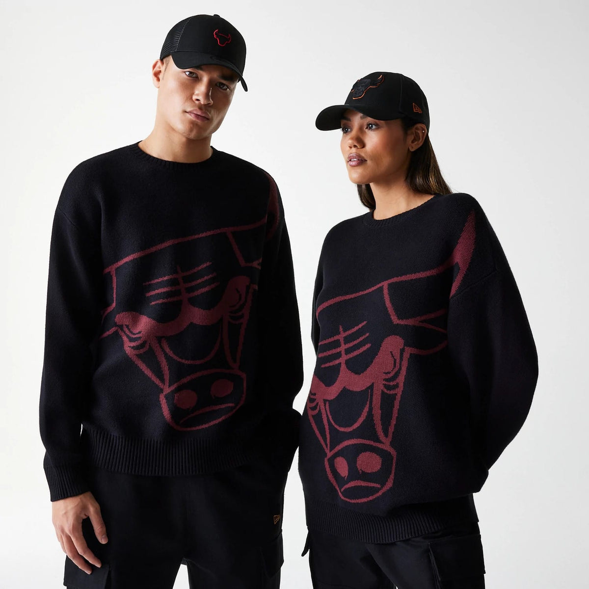 New Era Chicago Bulls NBA Knit Black Oversized Knit Sweater, on a model paired with a black Bull-logo cap, displaying the large red Bulls logo across the front, set against a black knitted fabric highlighting the oversized fit.
