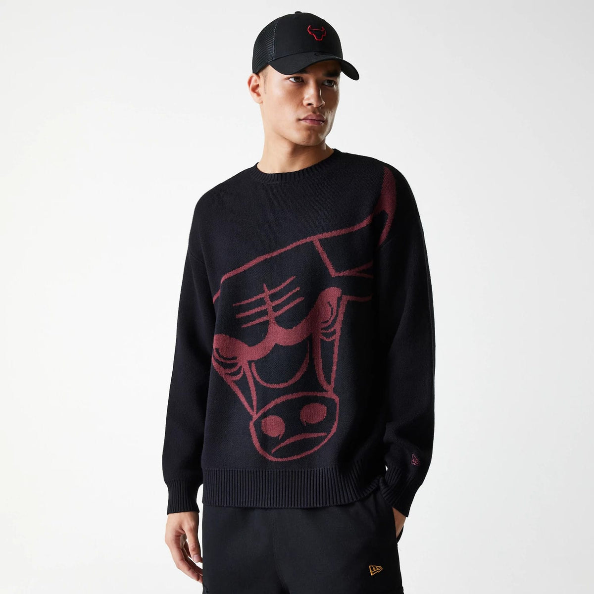 New Era Chicago Bulls NBA Knit Black Oversized Knit Sweater, front view on a model with a red Bulls logo cap, featuring an oversized red Bulls graphic knit on the front and a small embroidered New Era flag logo on the left cuff.