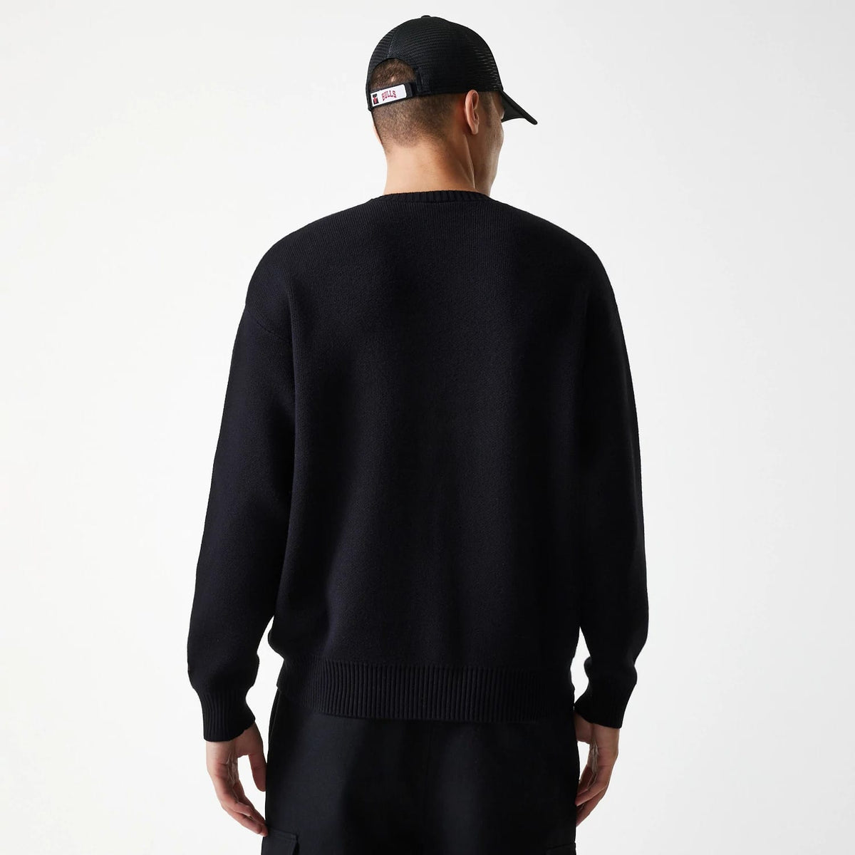 New Era Chicago Bulls NBA Knit Black Oversized Knit Sweater, rear view on a model with a black cap, showing the loose fit of the sweater and ribbed hem and cuffs, highlighting casual style with Chicago Bulls branding on the cap.