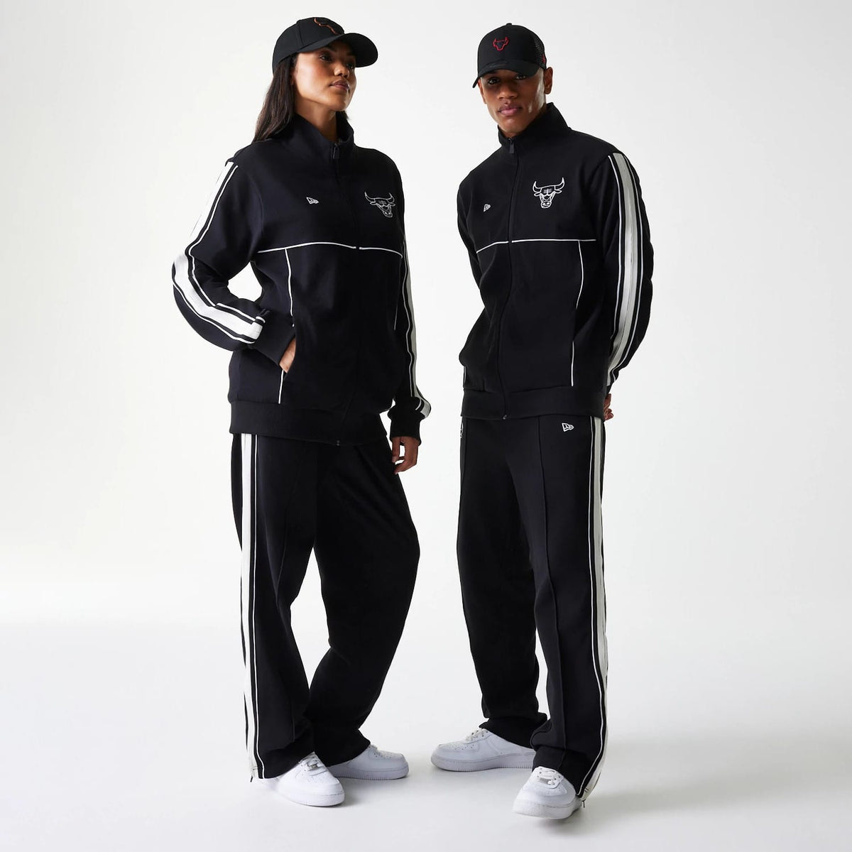 New Era Chicago Bulls NBA Outline Black Track Jacket, on-model view showing two individuals wearing black track jackets with a debossed Chicago Bulls logo on the chest, white piping along the sleeves, and matching black trousers with white stripes.