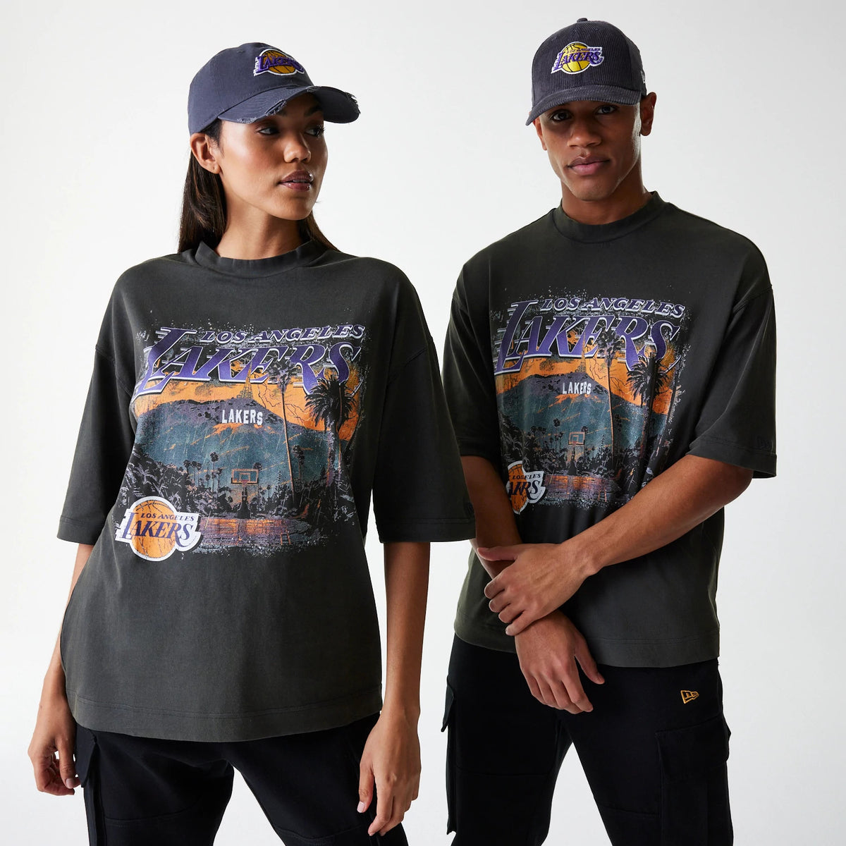 New Era LA Lakers NBA Washed Graphic Grey T-Shirt, on-model front view showing two people wearing dark grey tees with a large LA Lakers graphic in purple and orange on the chest, paired with caps featuring a raised LA Lakers logo.