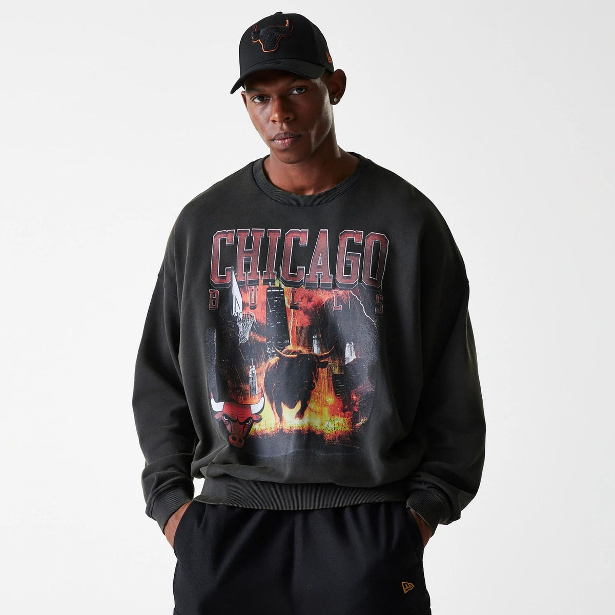 New Era Chicago Bulls NBA Washed Graphic Grey Oversized Crew Neck Sweater, front view on a model with a black cap, featuring a large red Chicago Bulls logo and graphic with a flaming bull and cityscape details.
