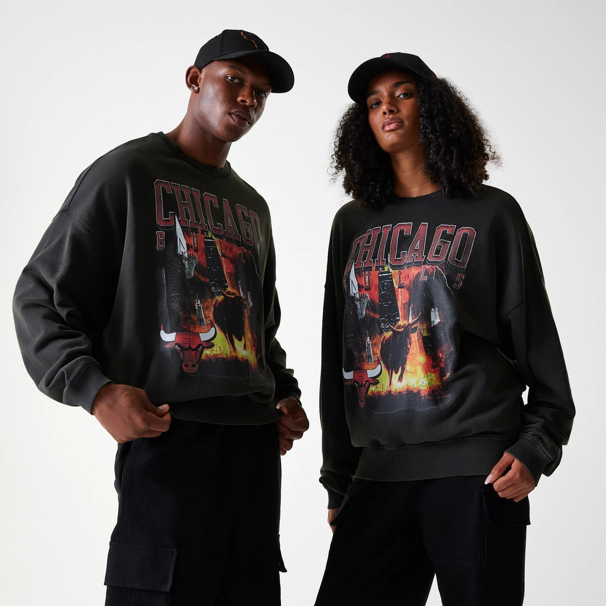 New Era Chicago Bulls NBA Washed Graphic Grey Oversized Crew Neck Sweater, on-model showing two individuals wearing matching black caps and sweaters featuring large Chicago Bulls text and graphic print on the front.