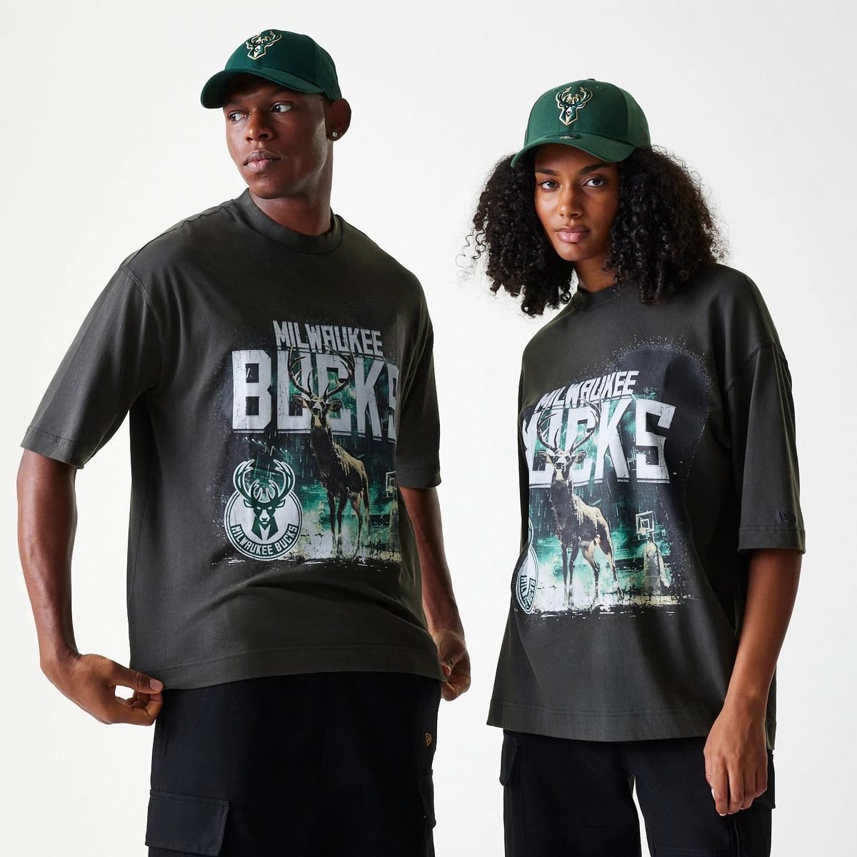 New Era Milwaukee Bucks NBA Washed Graphic Grey T-Shirt, on-model view showcasing large graphic print with team name and deer logo on the front, worn by models each styled with a dark green cap featuring an embroidered logo at the front.