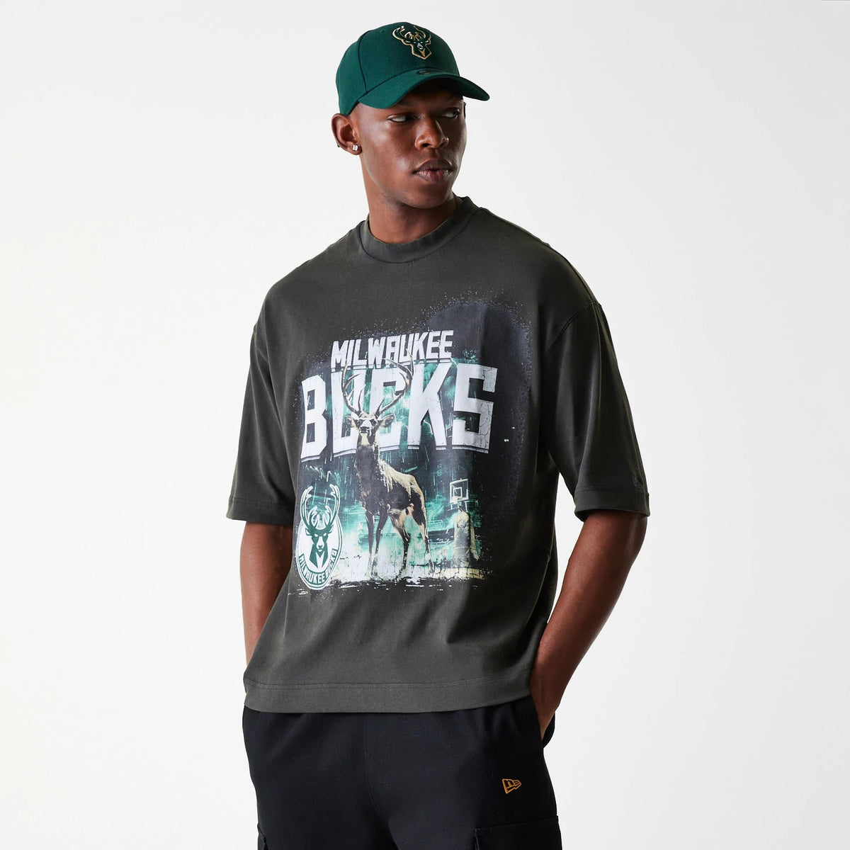 New Era Milwaukee Bucks NBA Washed Graphic Grey T-Shirt, on-model view showing the large graphic with BUCKS text and a deer image on the chest, paired with a green cap featuring a raised team logo on the front.
