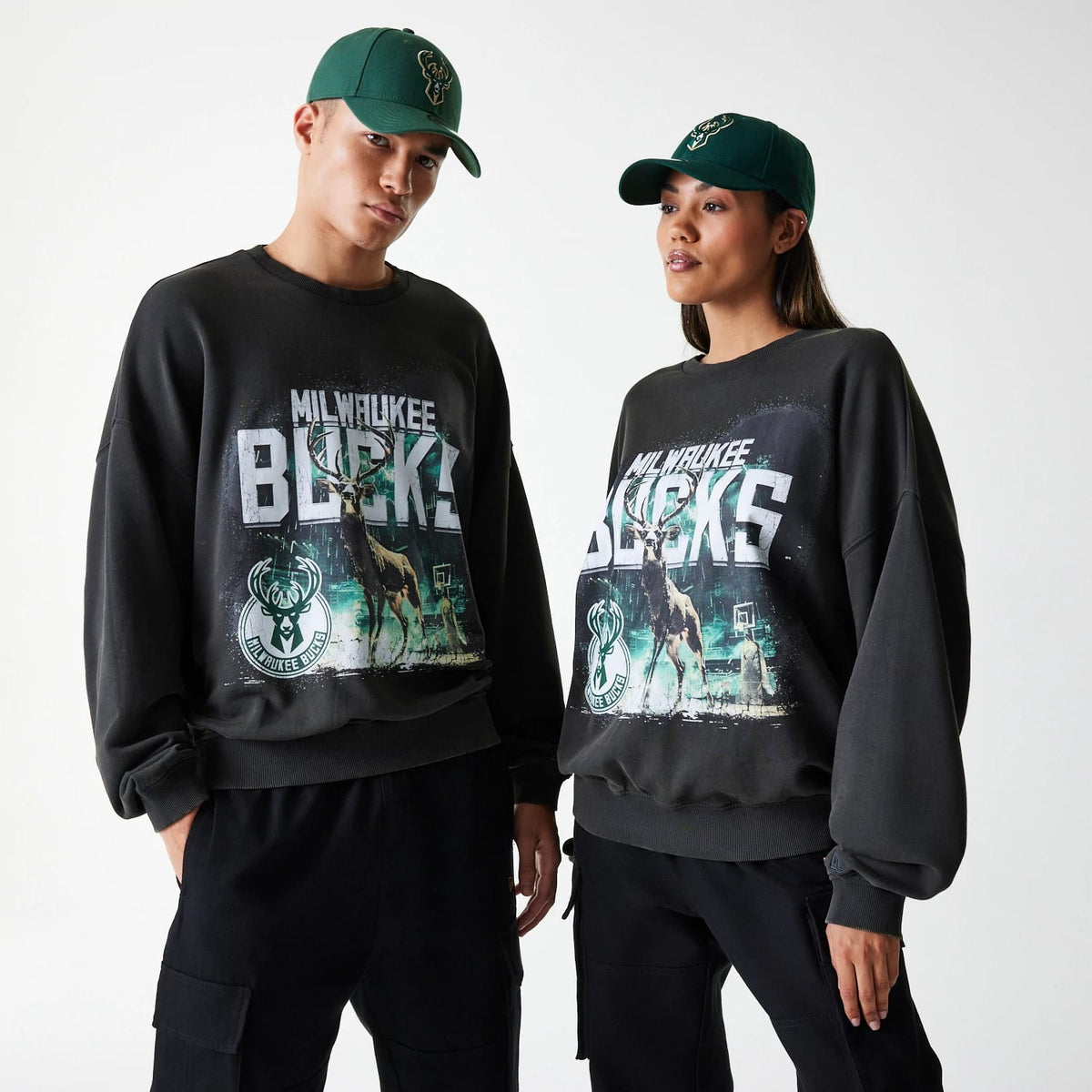 New Era Milwaukee Bucks NBA Washed Graphic Grey Oversized Crew Neck Sweater, front view on a male and female model wearing green Bucks caps, showcasing the large distressed Bucks lettering and mascot graphics on the chest.
