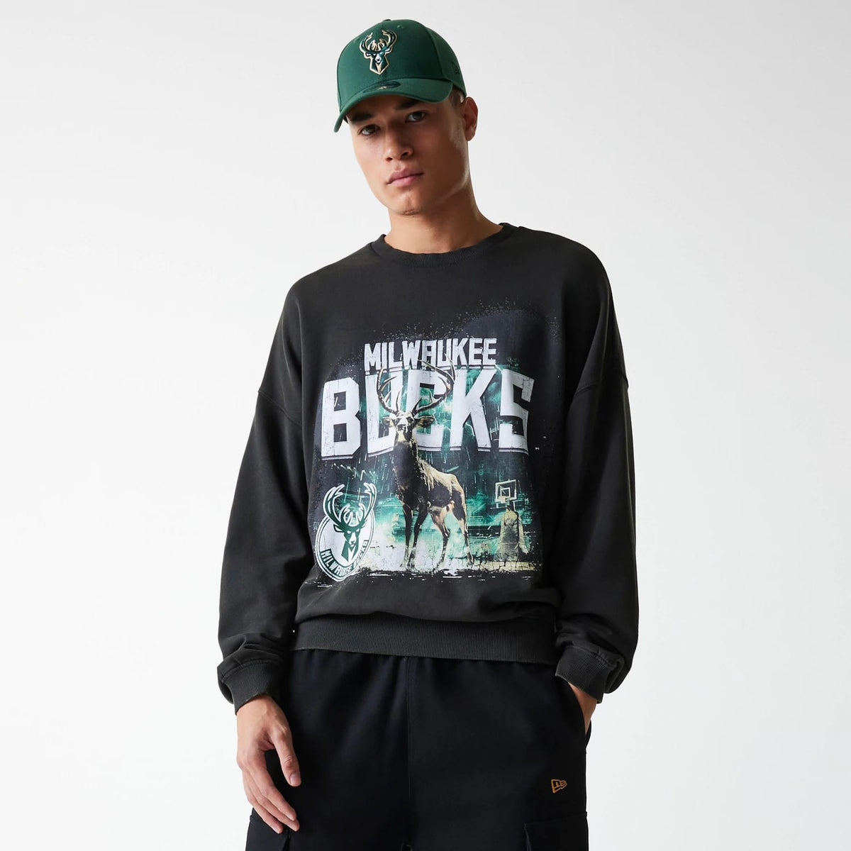 New Era Milwaukee Bucks NBA Washed Graphic Grey Oversized Crew Neck Sweater, on a model wearing matching dark trousers and a green Bucks cap, featuring a large Milwaukee Bucks graphic with a deer and basketball hoop on the front.