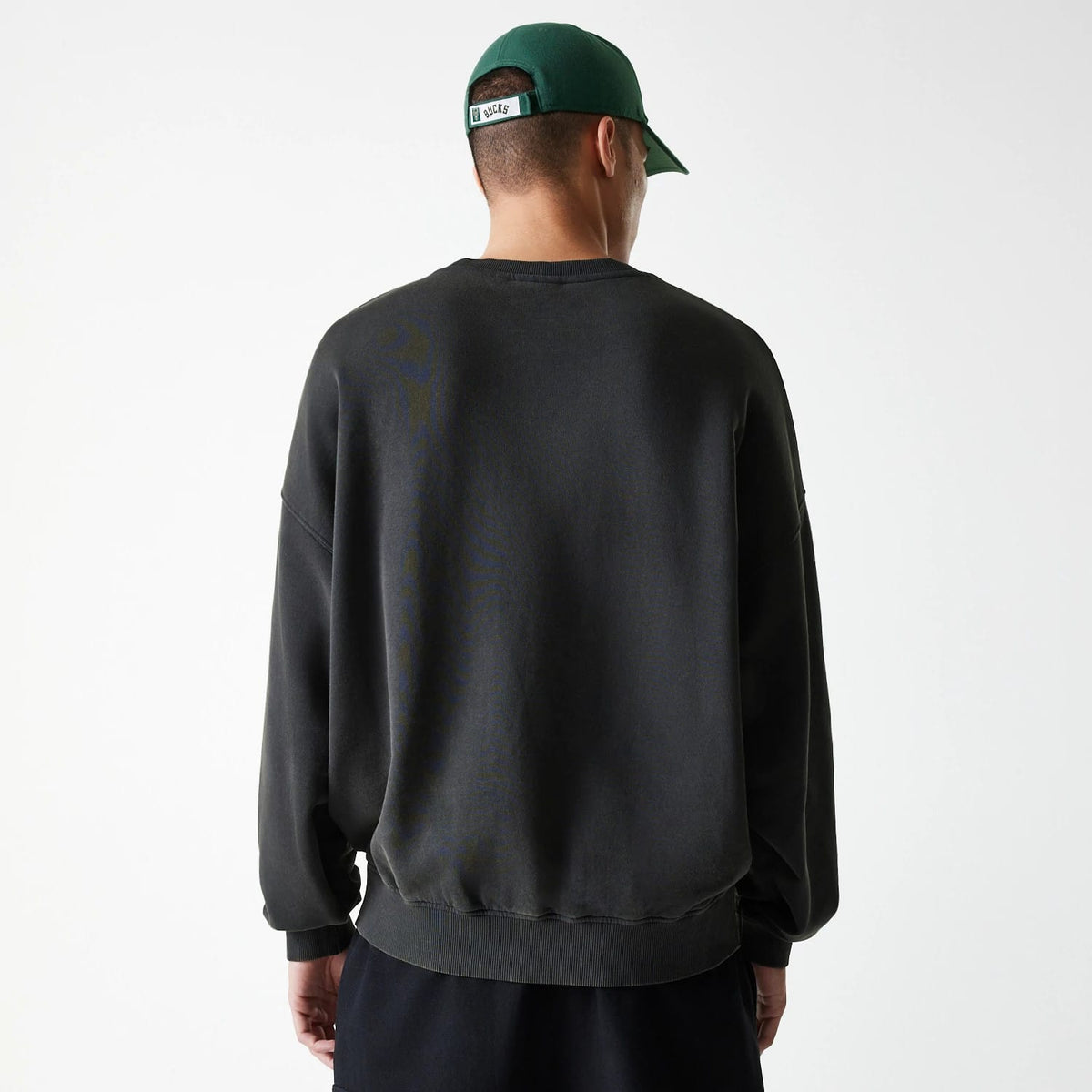 New Era Milwaukee Bucks NBA Washed Graphic Grey Oversized Crew Neck Sweater, back view on a model wearing a green cap, showing the relaxed fit and ribbed hem of the washed grey sweater.