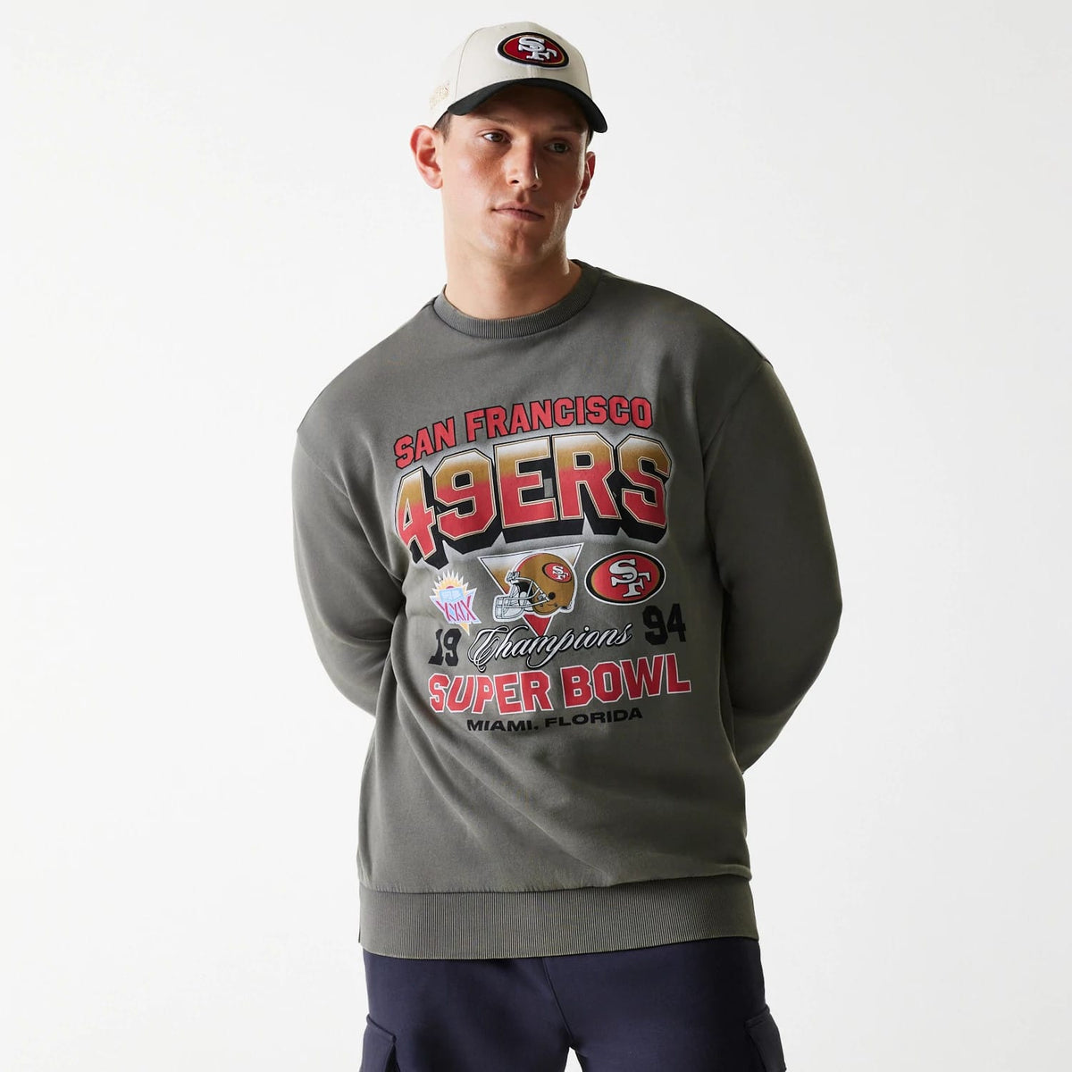 New Era San Francisco 49ers NFL Washed Super Bowl Grey Oversized Crew Neck Sweater, front view on a model with an off-white cap, featuring bold 49ERS lettering, championship and Super Bowl graphics in red, gold, and black across the chest.