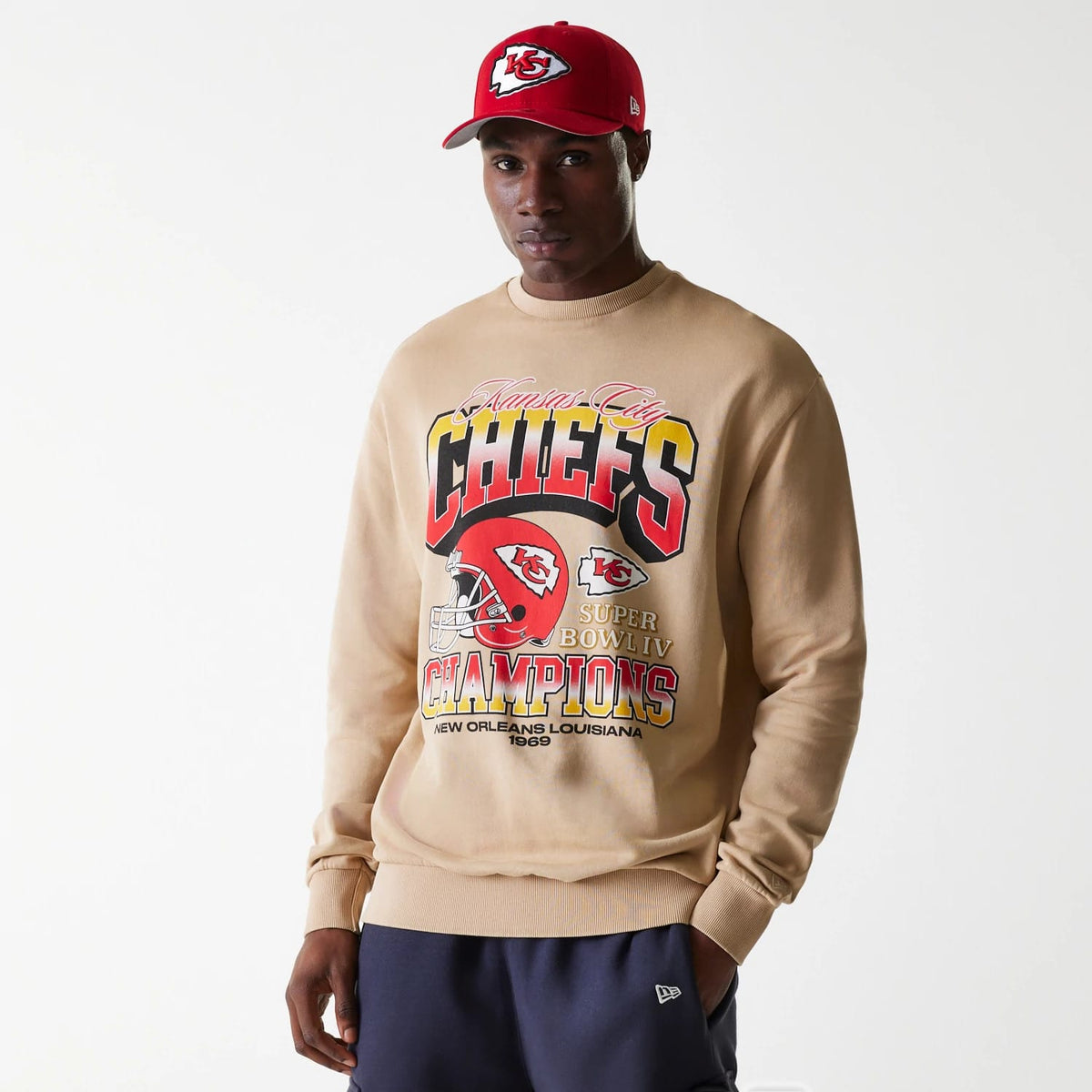 New Era Kansas City Chiefs NFL Washed Super Bowl Beige Oversized Crew Neck Sweater, front view on a model wearing a red cap with the Chiefs logo, featuring large Chiefs lettering and Super Bowl IV Champions graphics on the chest.