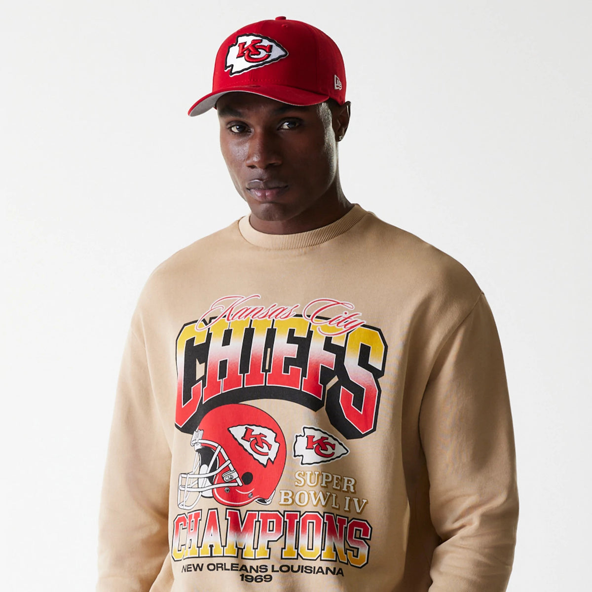 New Era Kansas City Chiefs NFL Washed Super Bowl Beige Oversized Crew Neck Sweater, on a model styled with a red cap featuring the Chiefs logo, showcasing bold yellow and red graphics with Super Bowl IV Champions text on the front.