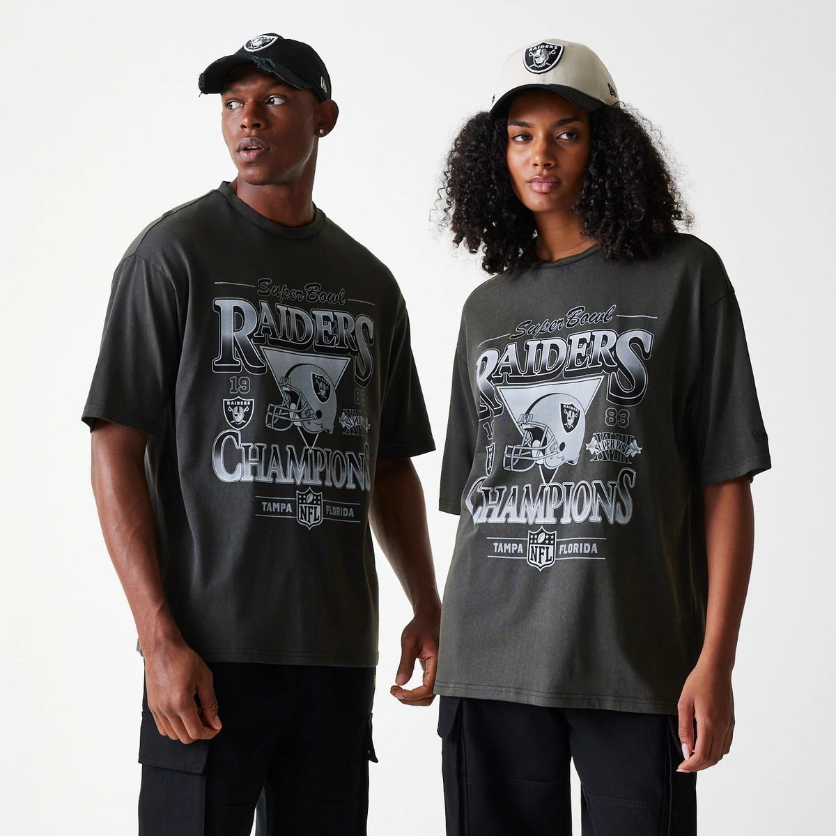 New Era Las Vegas Raiders NFL Washed Super Bowl Grey Oversized T-Shirt, on-model view showing two people wearing grey T-shirts with large front graphics featuring Raiders Champions text and a debossed logo, complemented by caps with debossed logos.