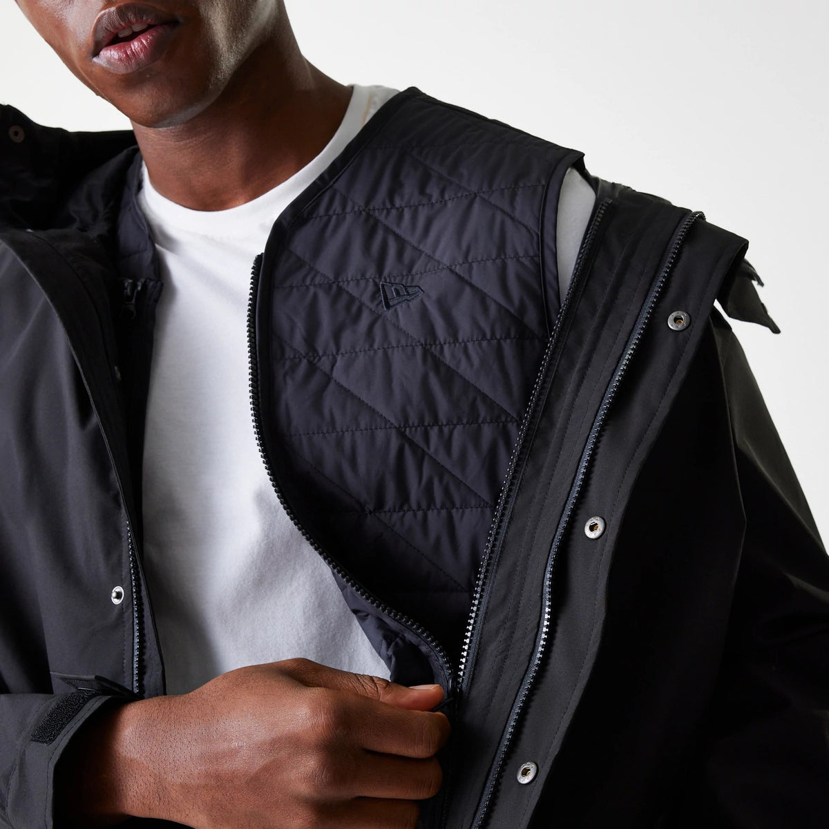 New Era Black Coach Jacket, front view showing the black quilted interior with a raised logo on the left side, a partially zipped closure with silver poppers along the edge and the jacket worn over a white shirt.