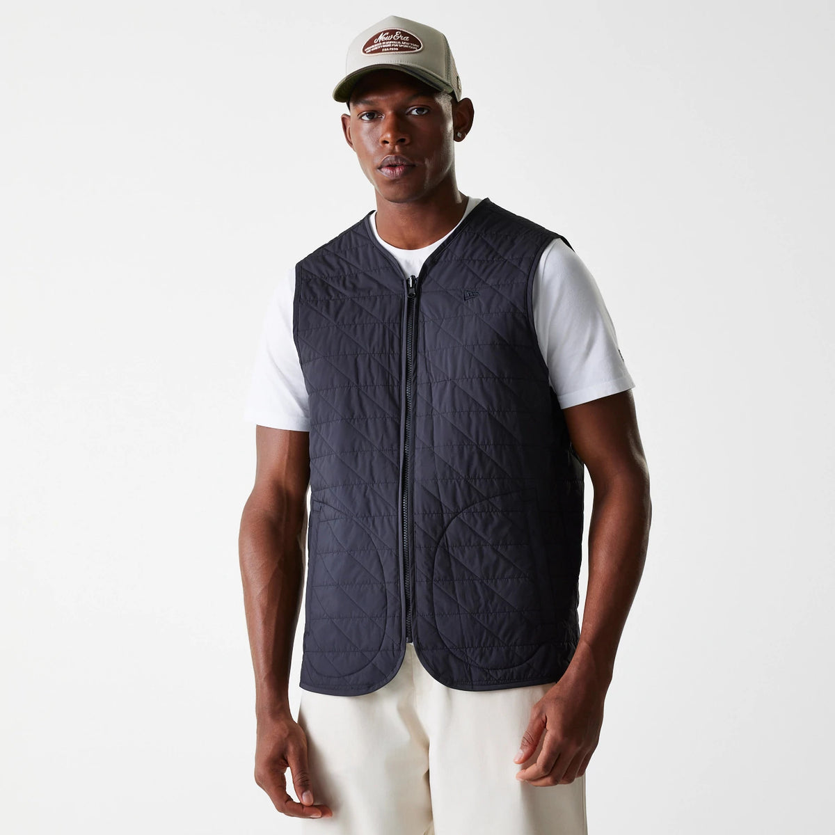 New Era Black Coach Jacket, on-model view showing the sleeveless black quilted jacket worn over a white T-shirt, with a central zip and a debossed logo on the left side of the chest.