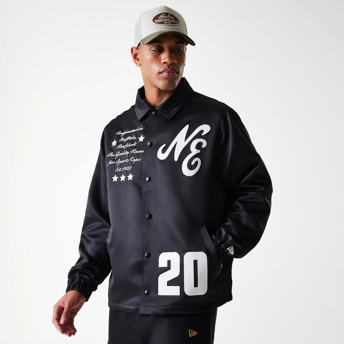 New Era Black Coaches Jacket, front view showing a black jacket with a large white NE and 20 printed on the front, embroidered script and stars on the left chest, worn by a model in a grey and brown cap.