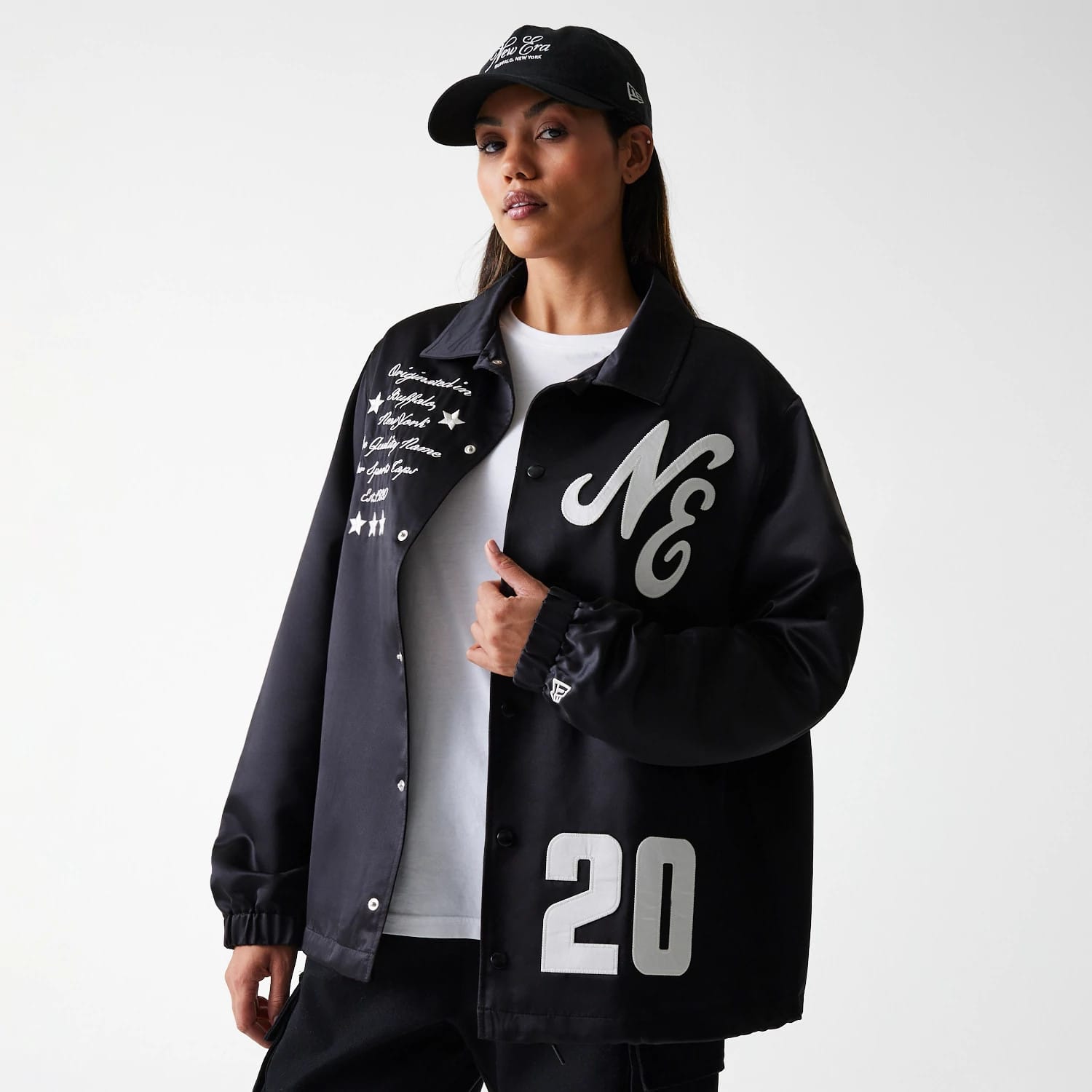 New Era Black Coaches Jacket, on-model view showing chest with a large 'NE' logo on the left side, embroidered script and stars on the right, and the number '20' at the hem, paired with a black cap.