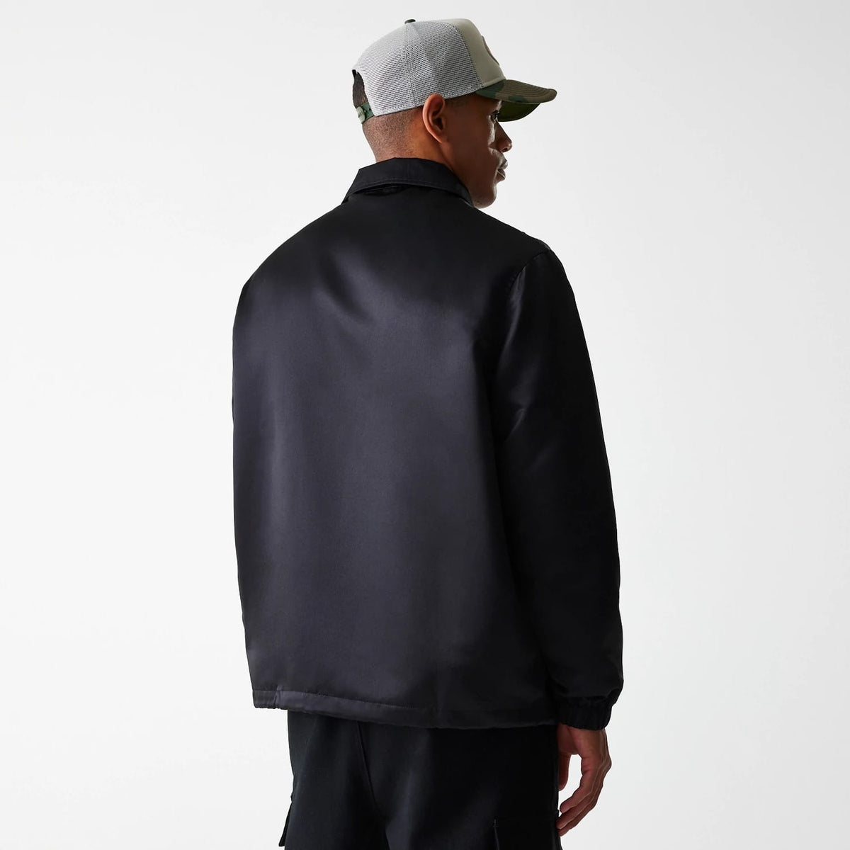 New Era Black Coaches Jacket, rear view showing the sleek black back panel extending from the collar down to the hem, paired with elasticated cuffs, worn by a person in a grey and camouflage cap.