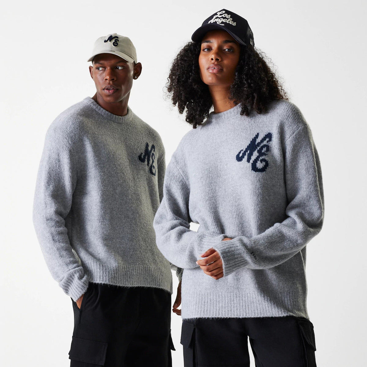 New Era Knit Grey Oversized Knit Sweater, on-model view showing two individuals wearing grey sweaters with navy NC logos on the chest, paired with black cargo trousers and New Era caps.
