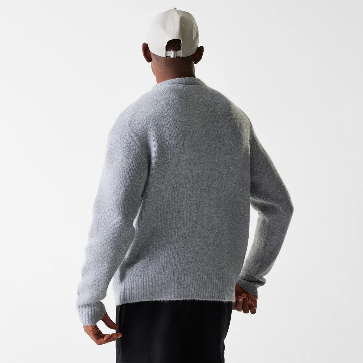 New Era Knit Grey Oversized Knit Sweater, back view on a model wearing a grey sweater with ribbed cuffs and hem, paired with black trousers and a cream cap featuring an adjustable buckle strap.
