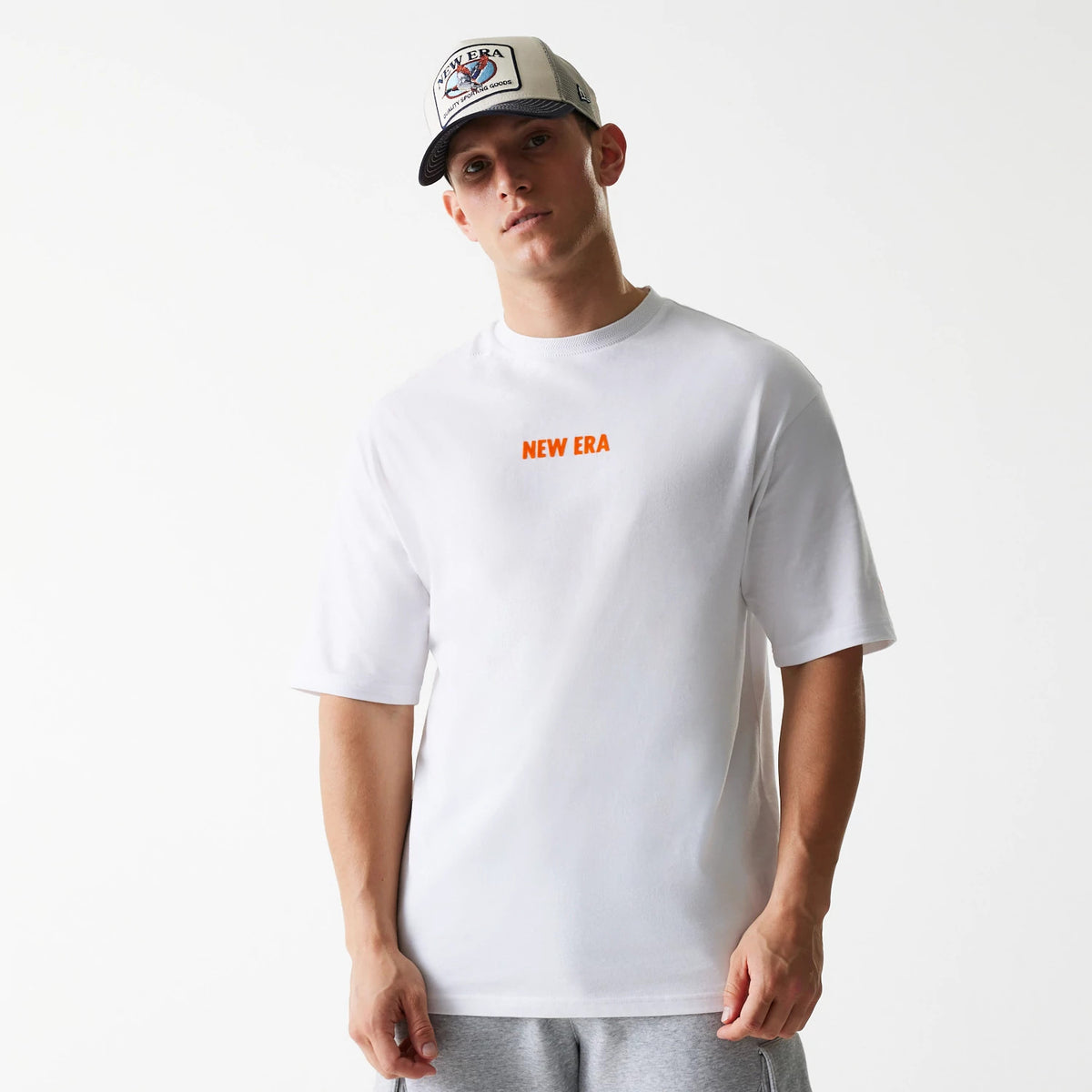 New Era Graphic White Oversized T-Shirt, front view showing the orange New Era logo centred on the chest, short sleeves, and a relaxed fit, worn by a model pairing it with a grey New Era cap featuring an embossed logo.