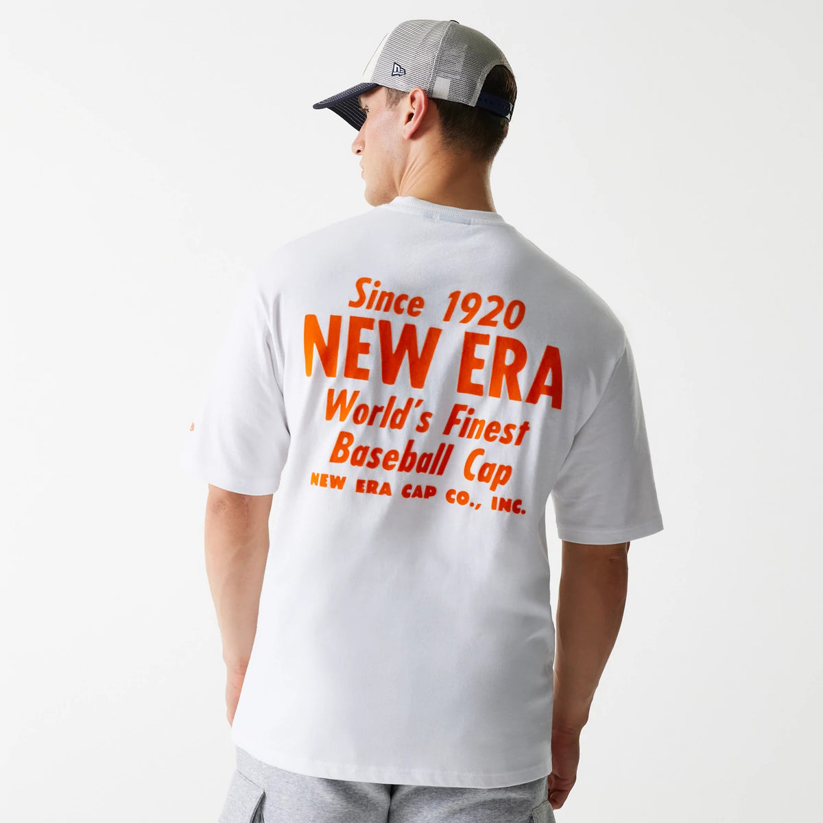 New Era Graphic White Oversized T-Shirt, rear view showing the large orange printed text Since 1920 NEW ERA World's Finest Baseball Cap centred on the white fabric, worn by a model in a grey and black cap.