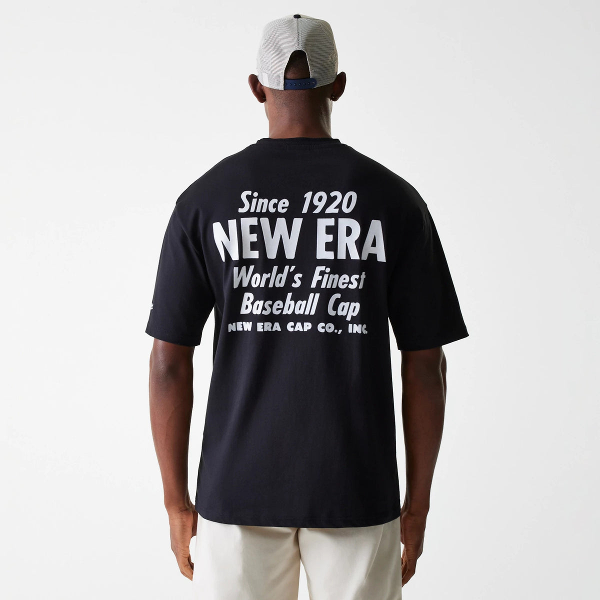 New Era Graphic Black Oversized T-Shirt, rear view showing large white graphic text stating Since 1920 NEW ERA World's Finest Baseball Cap across the back of the black T-shirt, worn by a person wearing a light cap.