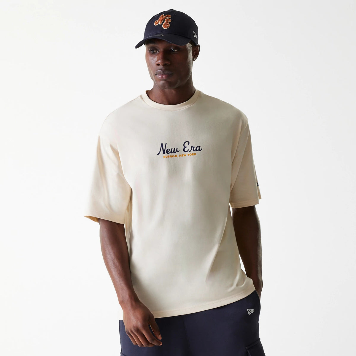 New Era Animal Graphic Light Beige Oversized T-Shirt, on-model view showing the light beige colour with New Era Buffalo New York text logo across the chest, paired with a black cap featuring an orange graphic logo.