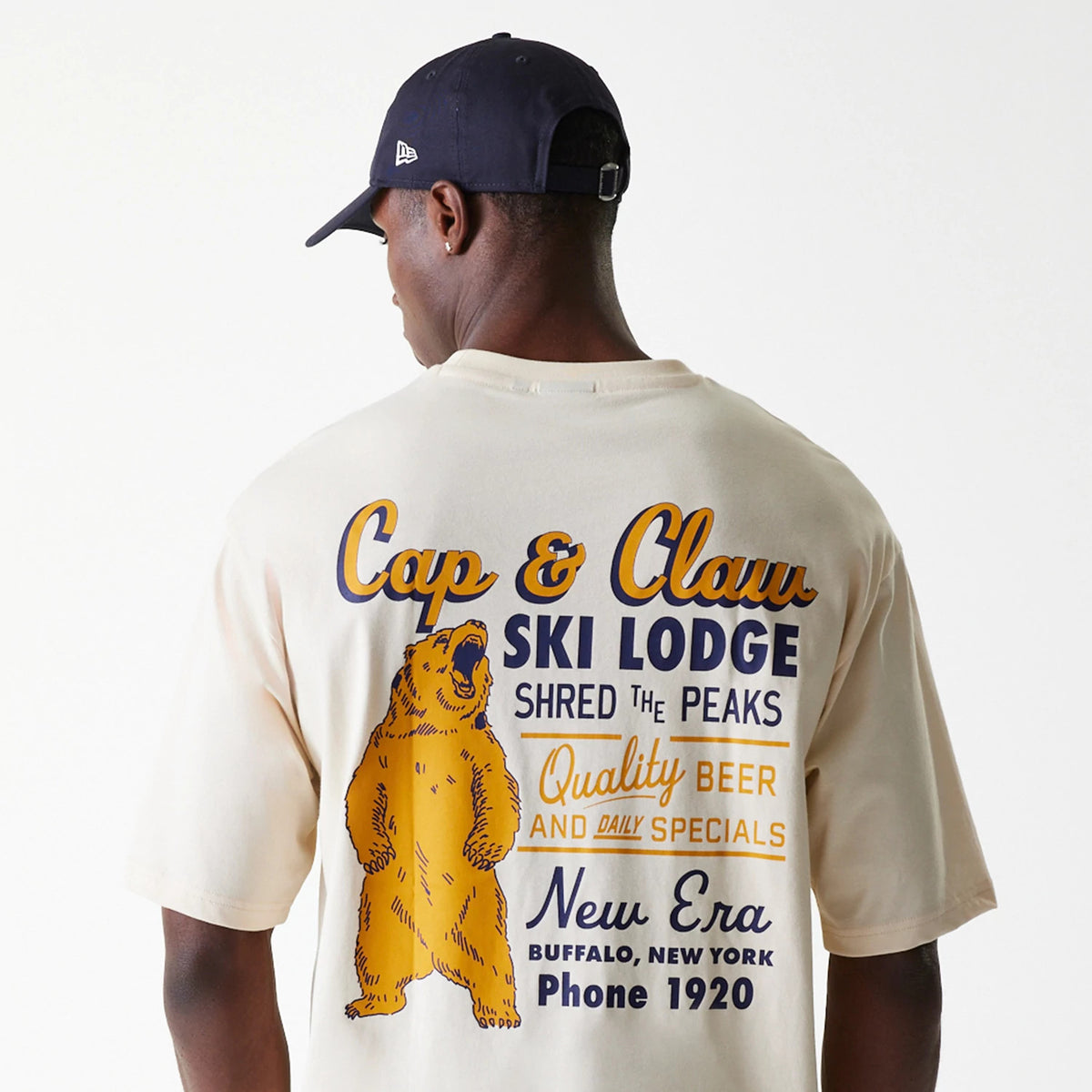 New Era Animal Graphic Light Beige Oversized T-Shirt, on-model back view showing the large colourful graphic with a bear and ski lodge text, worn by a person in a navy cap featuring a raised New Era logo on the left side.