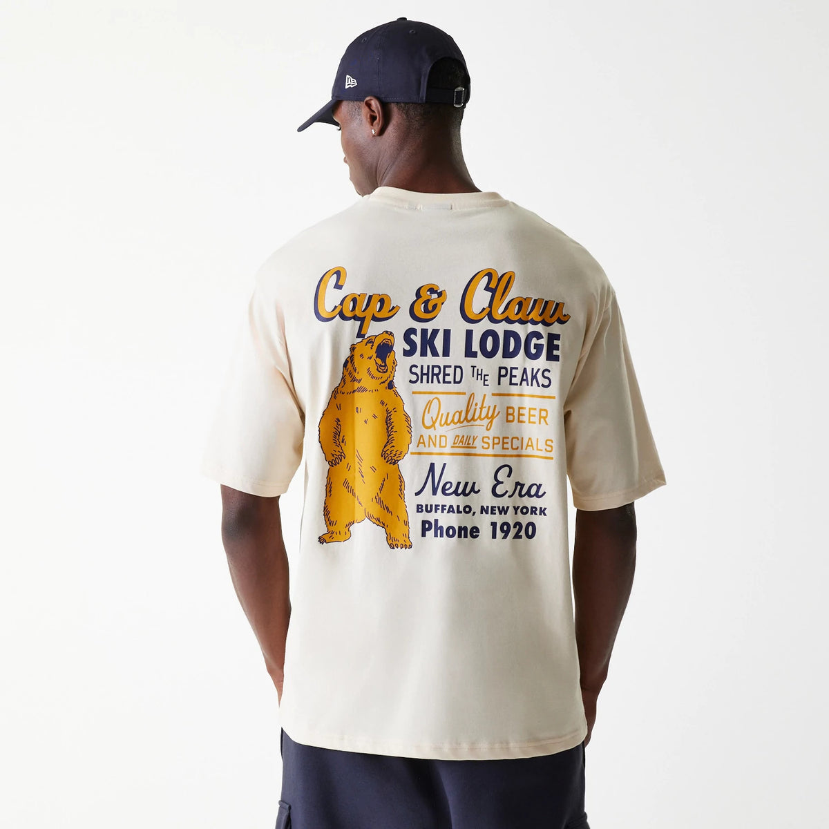 New Era Animal Graphic Light Beige Oversized T-Shirt, on-model view showing the rear with a large bear graphic and vintage lettering promoting a ski lodge, including slogans and a New Era logo, on a light beige background.