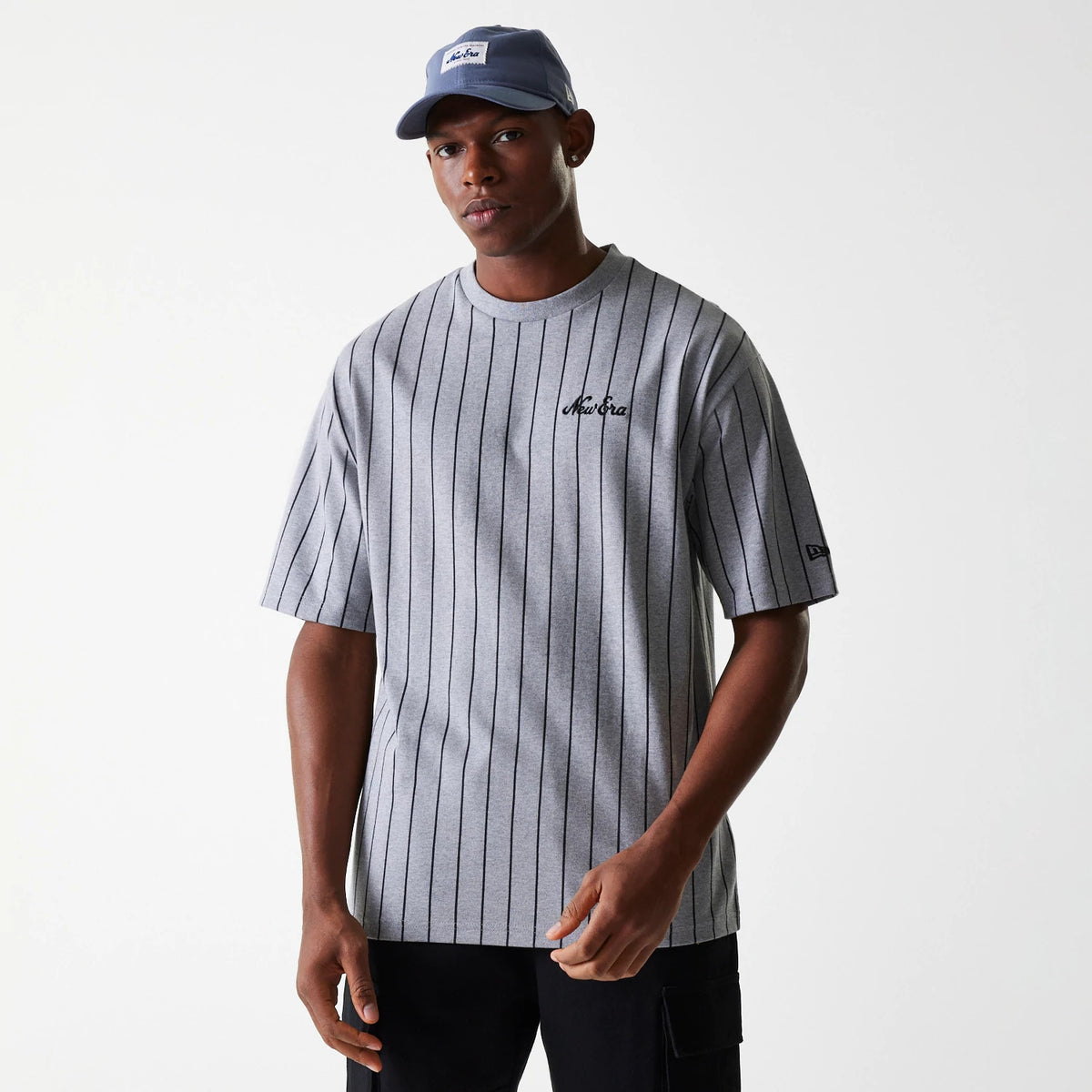 New Era Pinstripe Pastel Grey Oversized T-Shirt, on-model view showing the grey pinstriped shirt with a debossed New Era logo on the chest and a small New Era patch on the left sleeve, paired with a blue cap featuring a white patch.