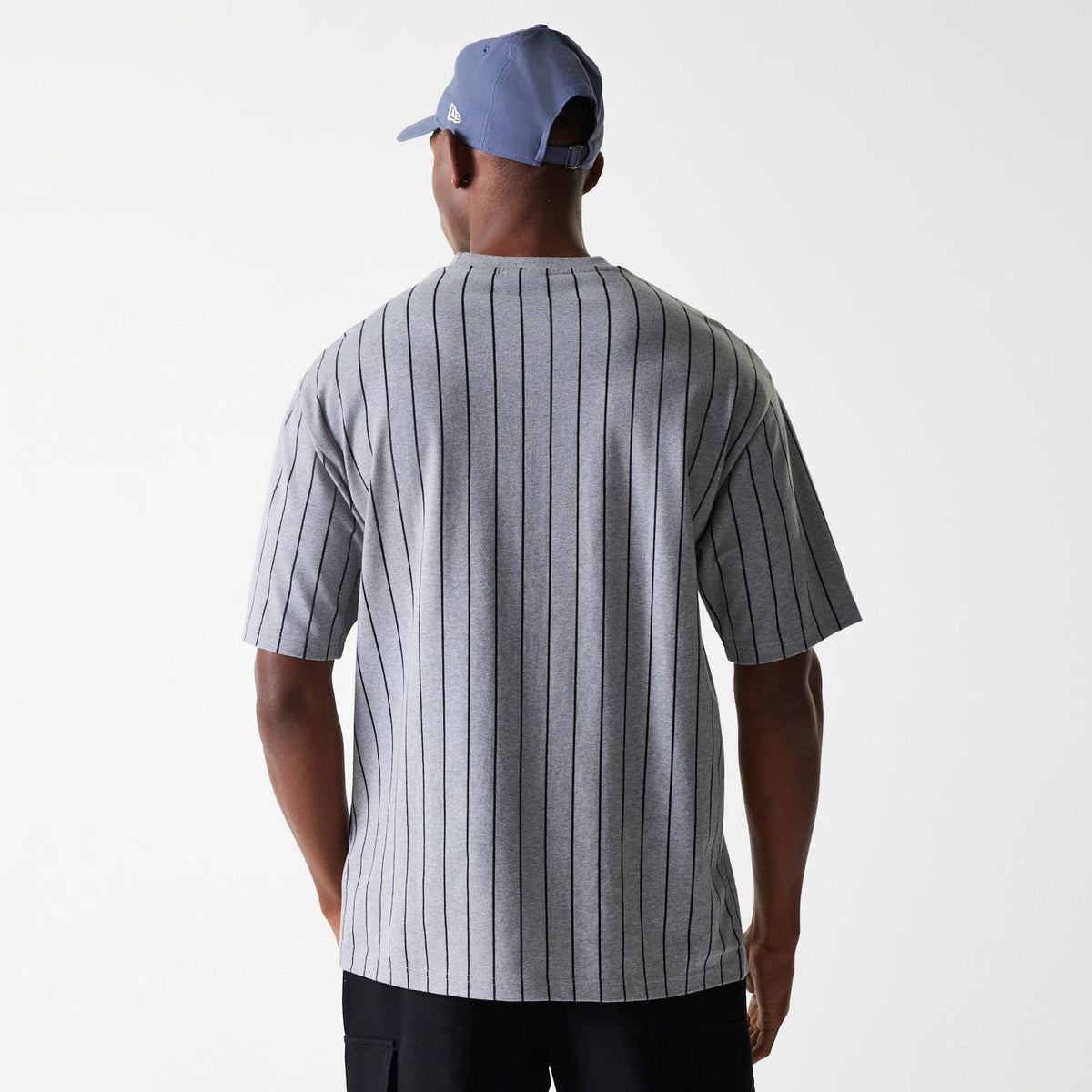 New Era Pinstripe Pastel Grey Oversized T-Shirt, on-model view showing the back with vertical black pinstripes on grey fabric, short sleeves and a crew neckline, paired with a man wearing a purple cap featuring a raised New Era logo on the side.