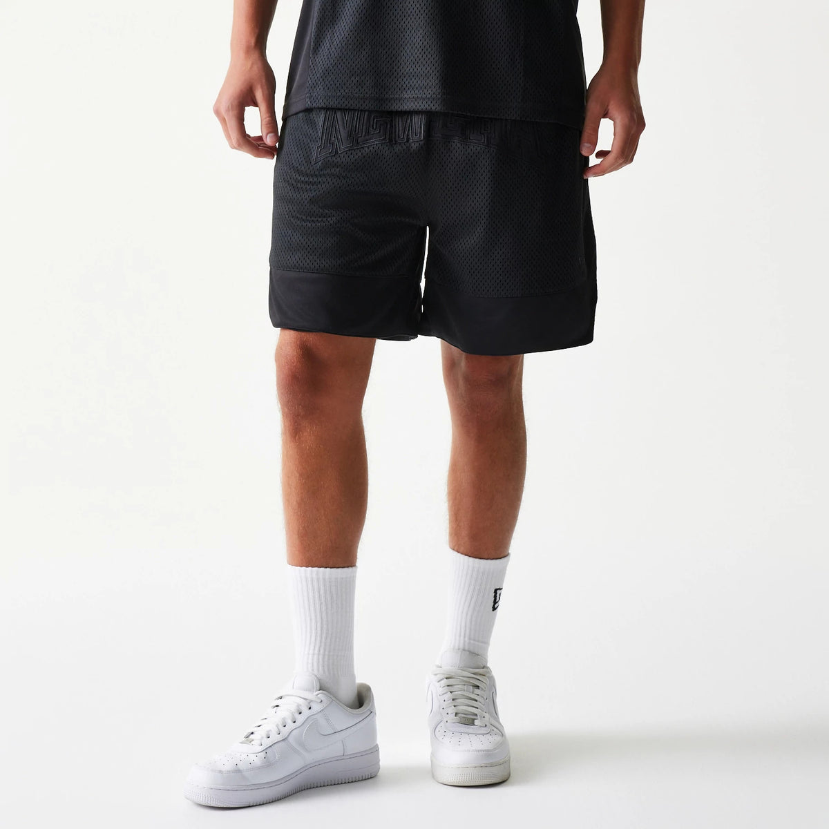New Era Mesh Black Shorts, on-model view showing the knee-length black mesh fabric with a raised logo across the elasticated waistband and solid black panels at the hems, worn with white sneakers and socks.