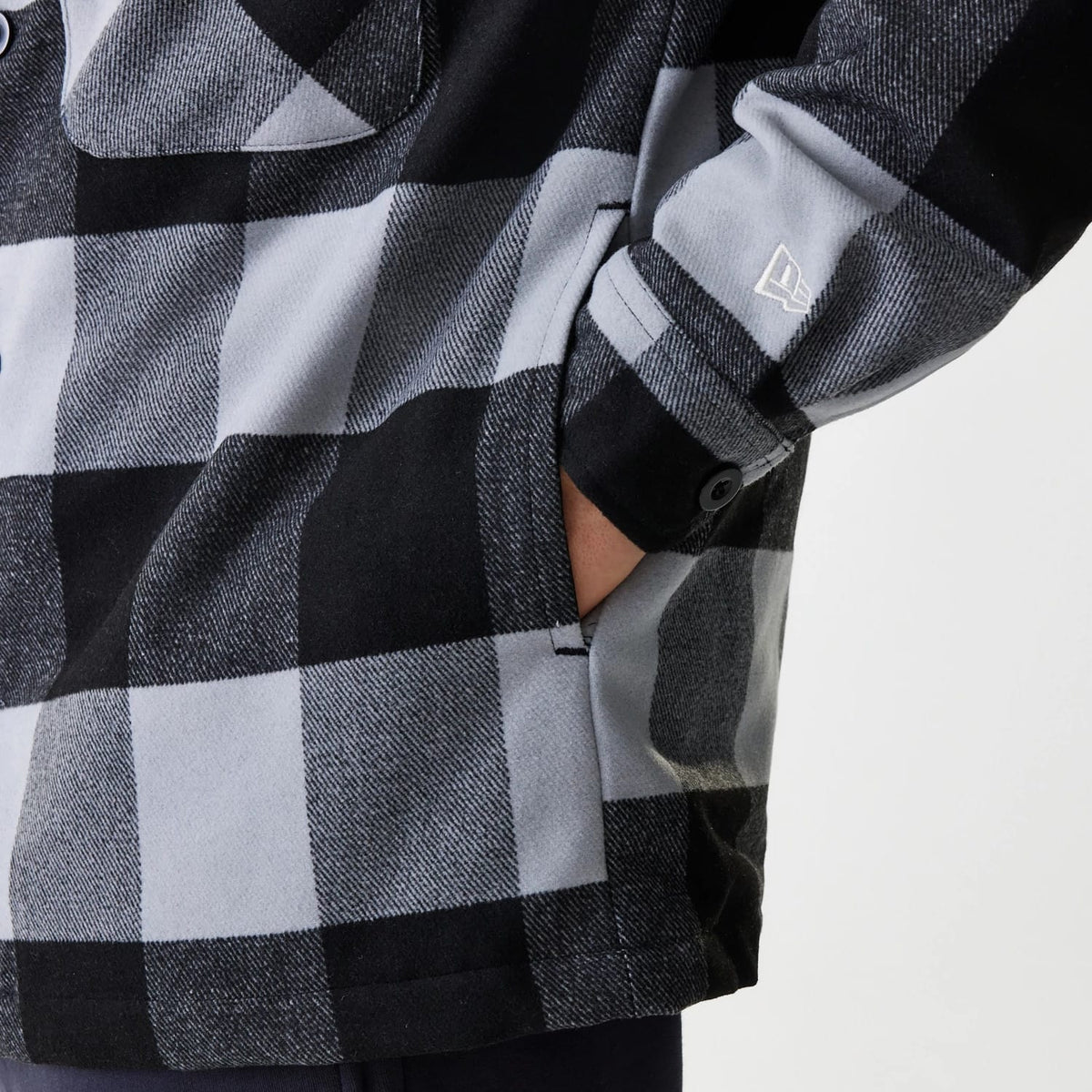 New Era Black Shacket, close-up showing black and grey check pattern, side pocket with a hand inside, black buttoned sleeve cuff, and New Era flag logo embroidered in white on the left sleeve.