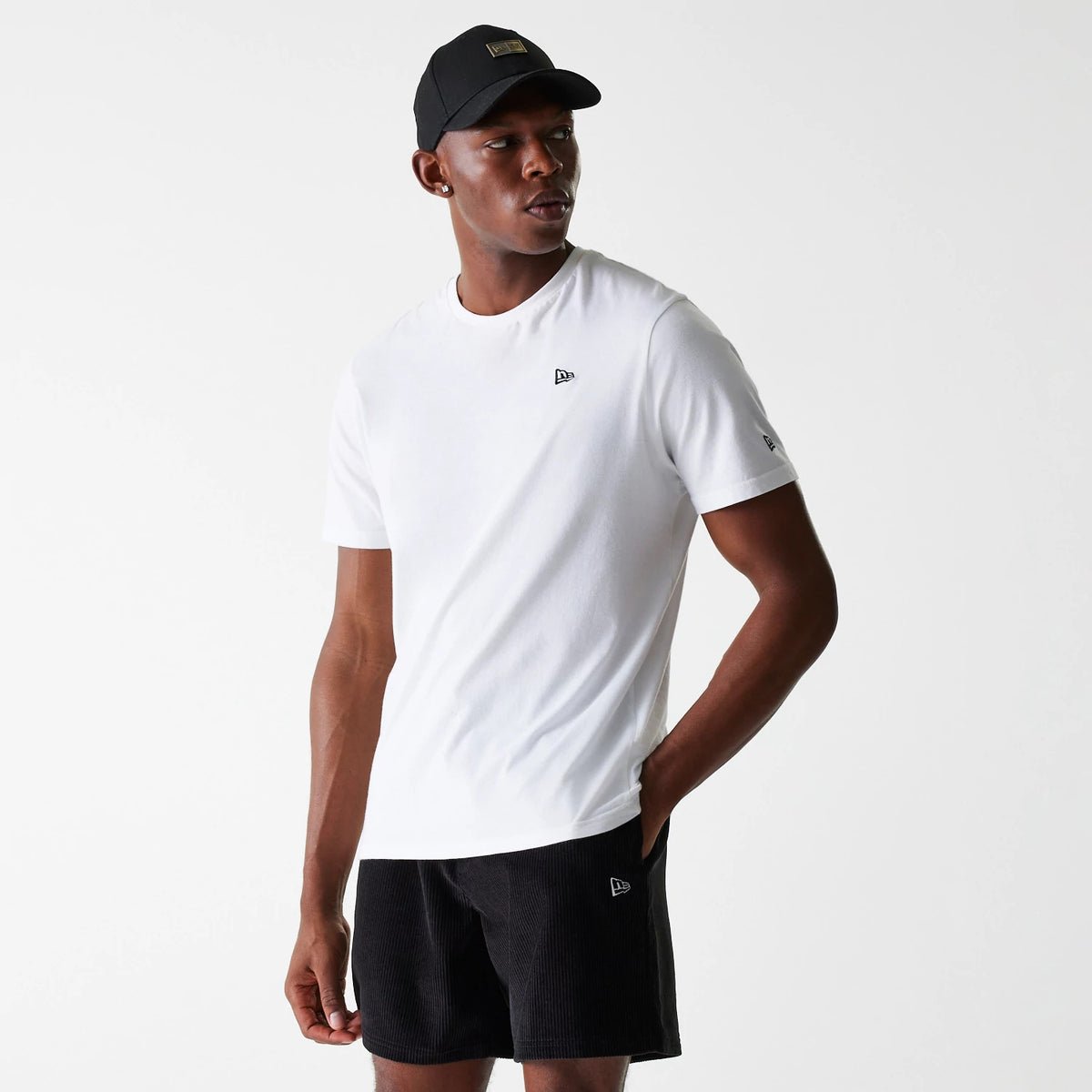 New Era Cord Black Shorts, on-model view showing the black corded shorts with an embroidered New Era logo above the left hem, paired with a white T-shirt featuring a small logo on the chest and a black cap with a patch logo.