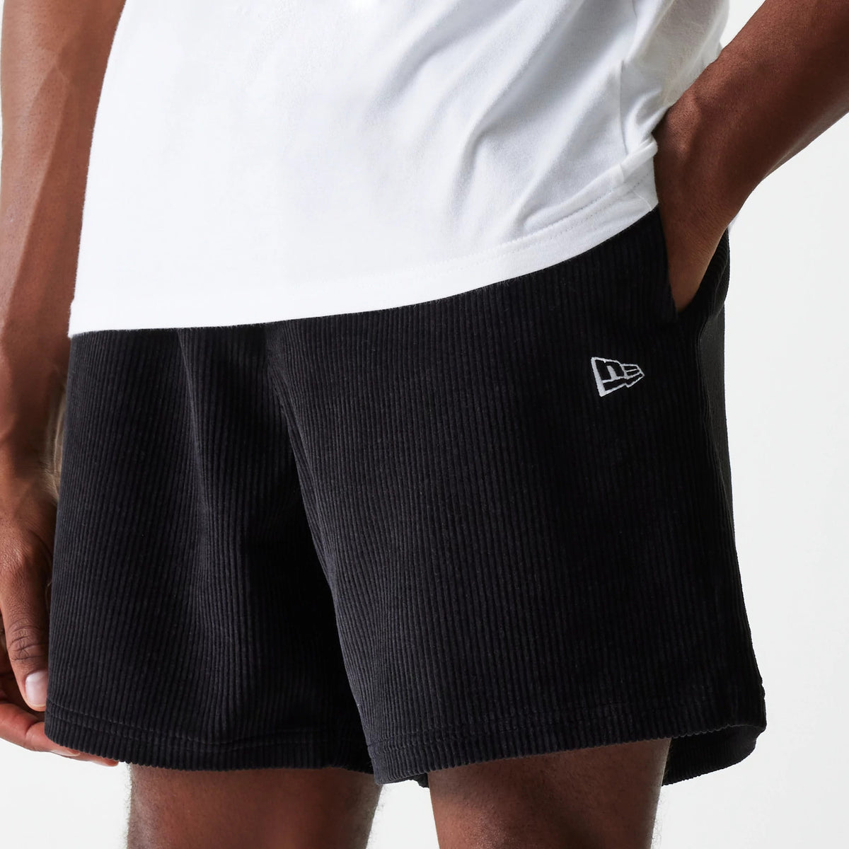 New Era Cord Black Shorts, three-quarter view showing the ribbed texture of the black shorts with a raised New Era logo on the lower left leg, worn by a model paired with a white top.
