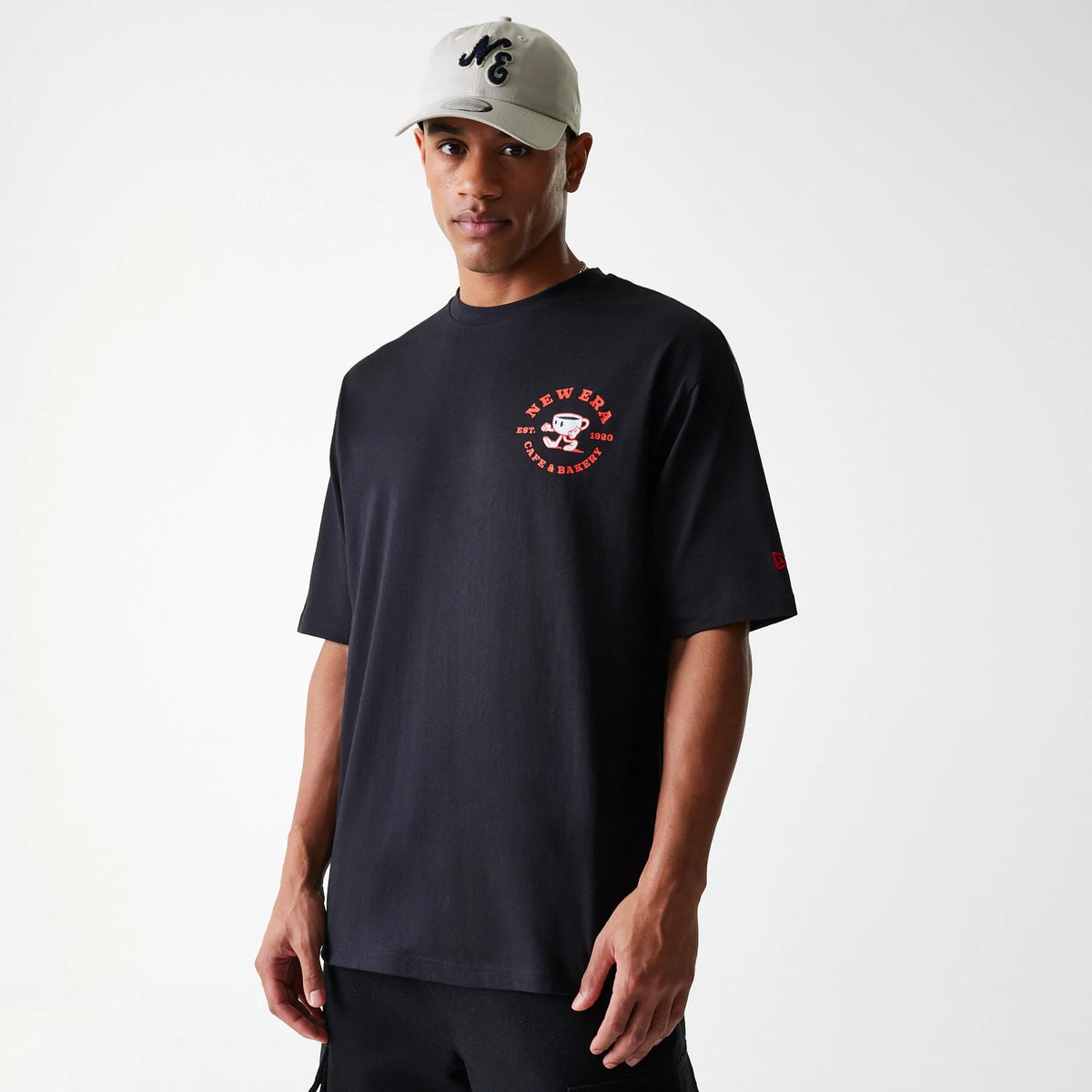 New Era Cafe Graphic Black Oversized T-Shirt, on-model view showing a black t-shirt with a red debossed New Era Cafe graphic logo on the left chest, styled with short sleeves and a crew neck, worn by a person in black shorts and a grey cap.