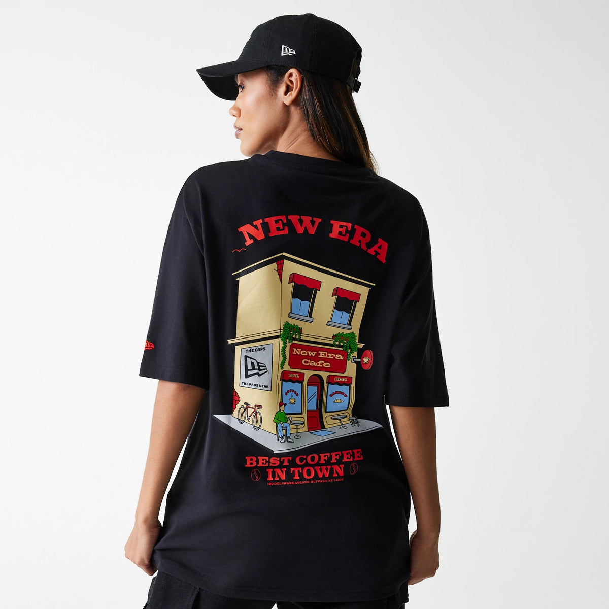 New Era Cafe Graphic Black Oversized T-Shirt, on-model view showing the large colourful graphic of a cafe on the back with red text reading New Era and Best Coffee in Town, paired with a raised New Era logo on a black cap.