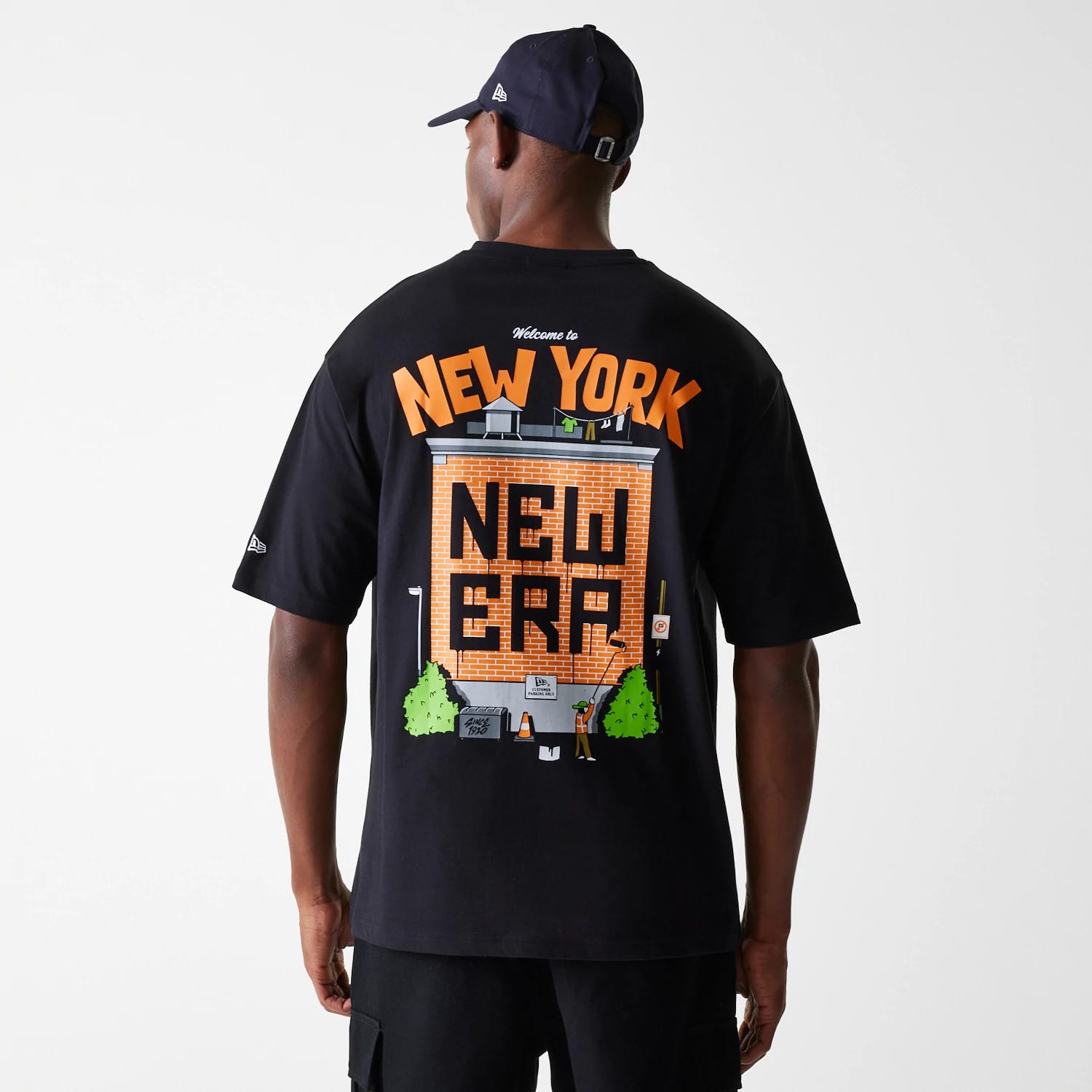 New Era Graffiti Graphic Black Oversized T-Shirt, rear view showing a large graphic of a brick building with New Era and Welcome to New York text in orange, worn by a person wearing a black cap with a debossed logo on the side.