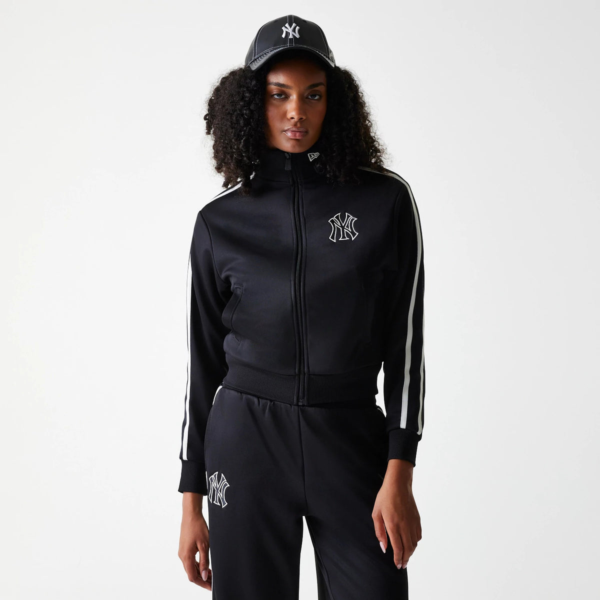 New Era New York Yankees Womens MLB Midi Logo Black Track Jacket, on-model view showing the black track jacket with a front zip and white stripes down the sleeves, the raised Yankees logo on the chest, and a raised New Era logo on a black cap worn.