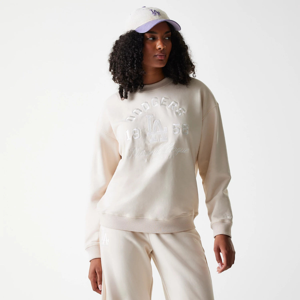New Era LA Dodgers Womens MLB Tonal Graphic Light Beige Oversized Crew Neck Sweater, front view on a model with matching beige trousers and a white cap, showing raised white Dodgers lettering and LA logo embroidery on the chest.