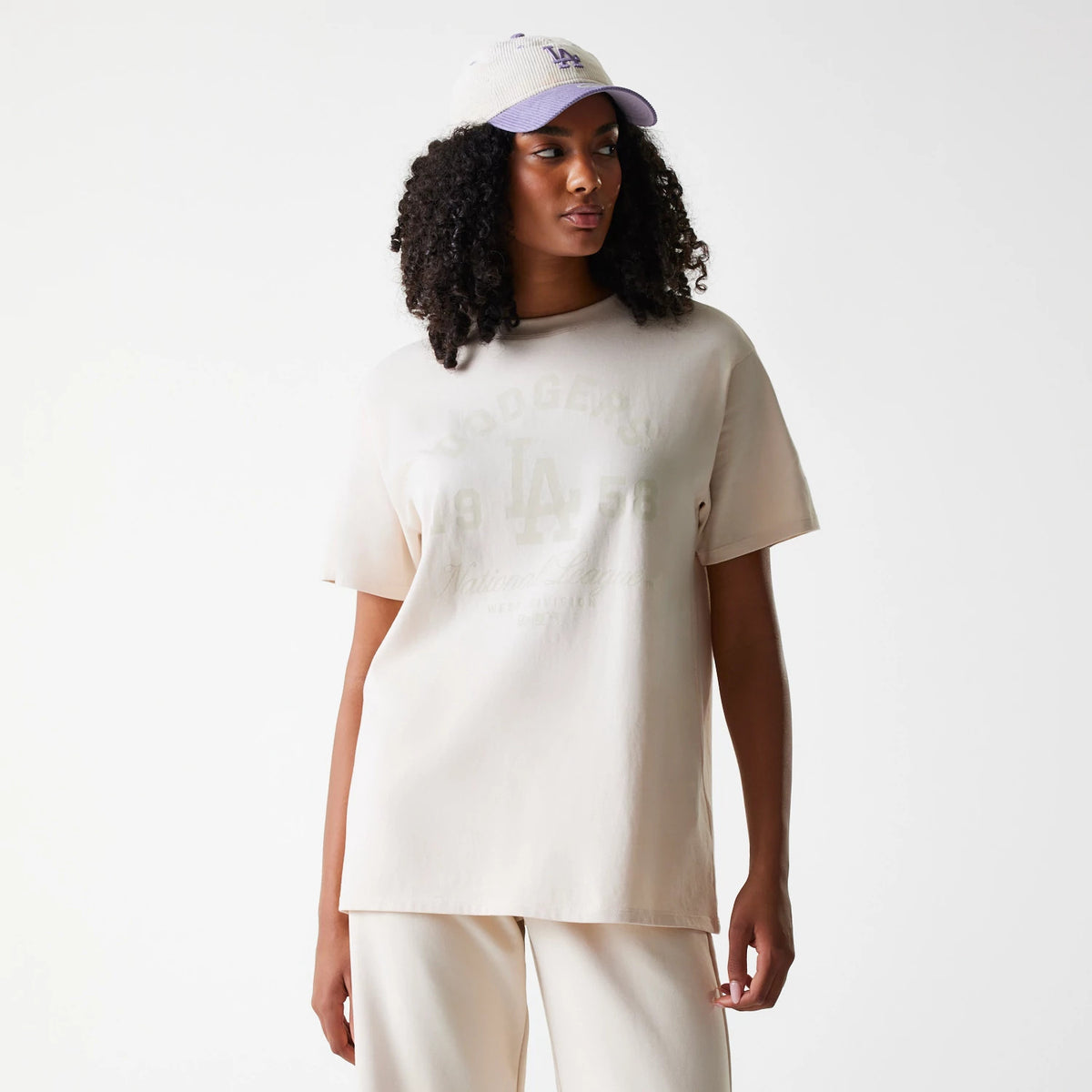 New Era LA Dodgers Womens MLB Tonal Graphic Light Beige Oversized T-Shirt, front view showing a tonal LA Dodgers graphic on the chest with short sleeves and a relaxed fit, paired with an on-model white cap featuring a raised purple LA logo.