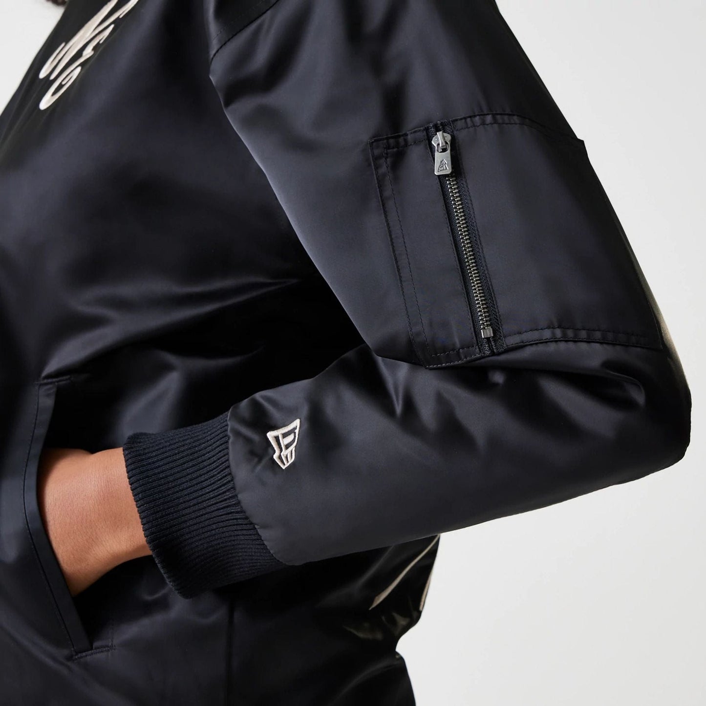 Womens New Era Black Satin Bomber Jacket, side view showing the sleek black sleeve with a zip pocket on the upper arm, ribbed cuff, and an embroidered New Era logo near the wrist.