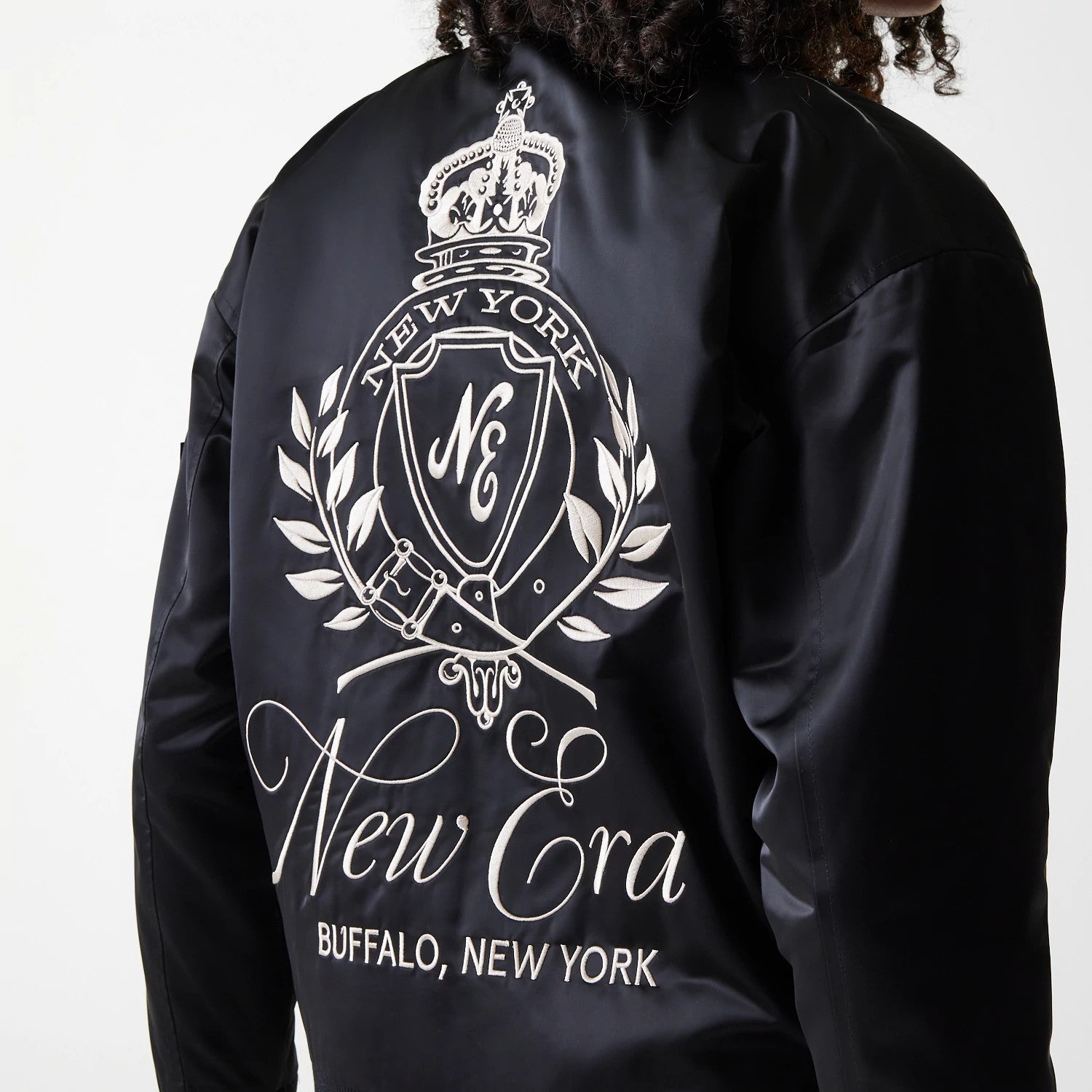 Womens New Era Black Satin Bomber Jacket, back view showing a large white embroidered crest design with New York and New Era text, along with flourishes and Buffalo, New York at the bottom of the black satin fabric.