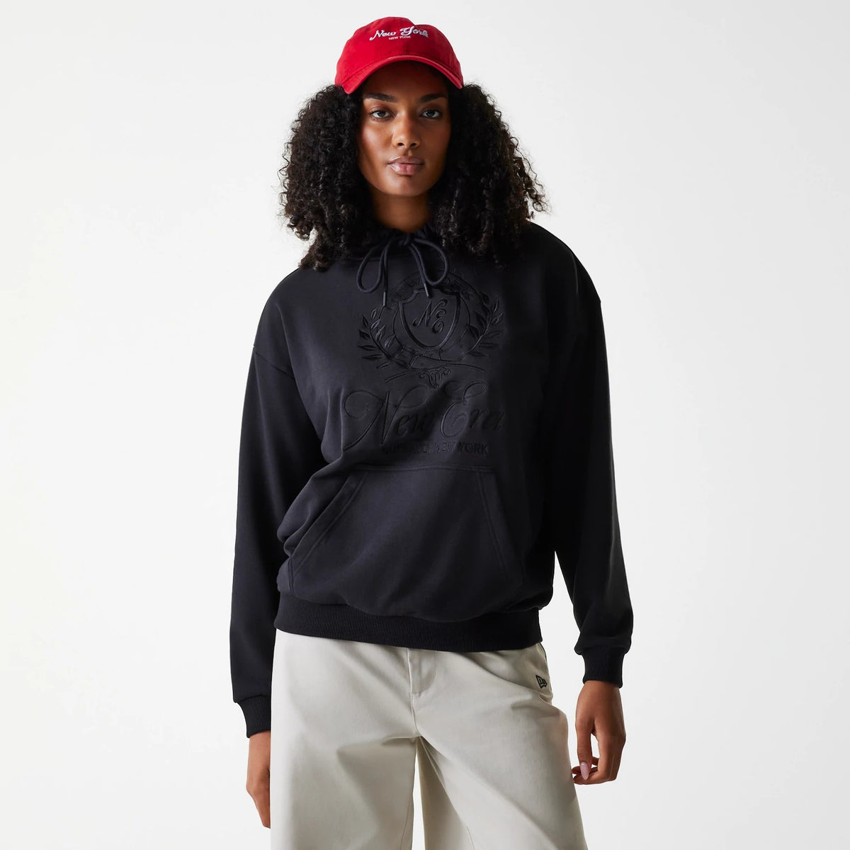 Womens New Era Crest Black Pullover Hoodie, front view showing the black hoodie with a large embossed crest and New Era logo on the chest, drawstrings hanging from the hood and a kangaroo pocket at the front, worn by a model with a red cap.