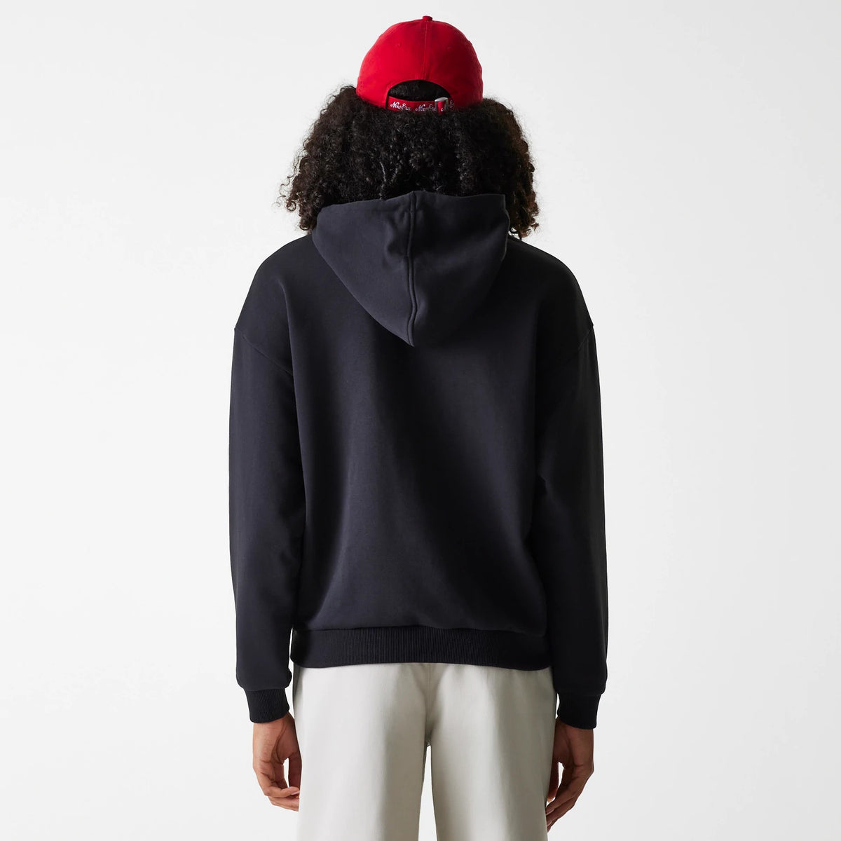 Womens New Era Crest Black Pullover Hoodie, rear view showing the relaxed black hoodie with a generous hood, the hem and cuffs ribbed for fit, worn by a model with a red cap, showcasing a casual look.
