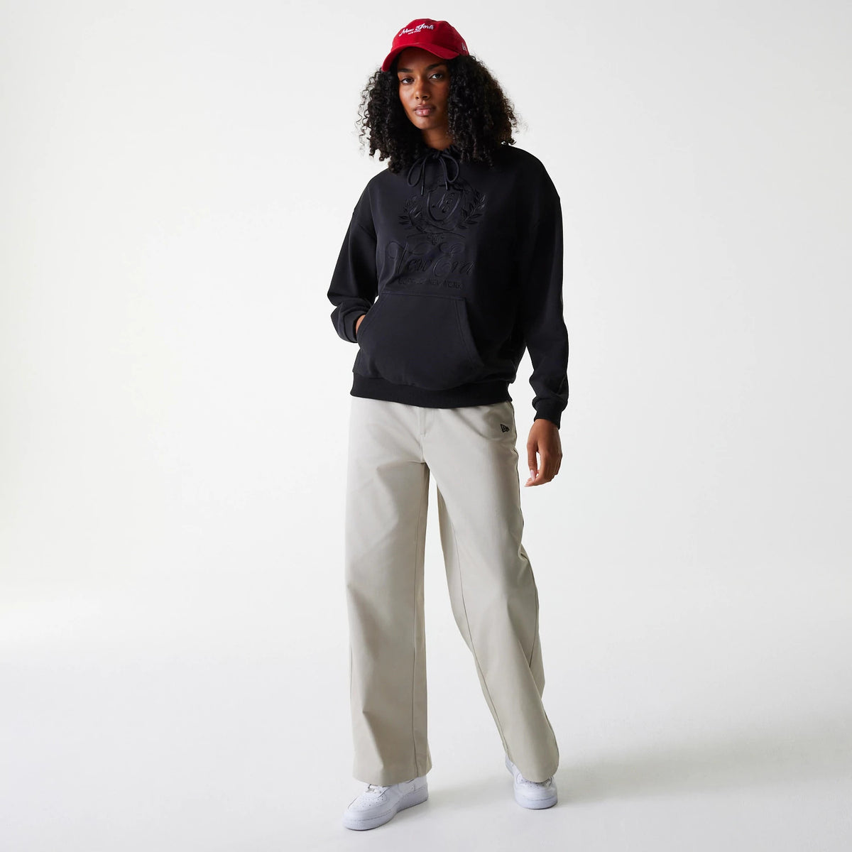 Womens New Era Crest Black Pullover Hoodie, on-model view showing a black hoodie with a large embossed crest logo on the chest, paired with beige trousers and a red cap featuring a white embossed logo.
